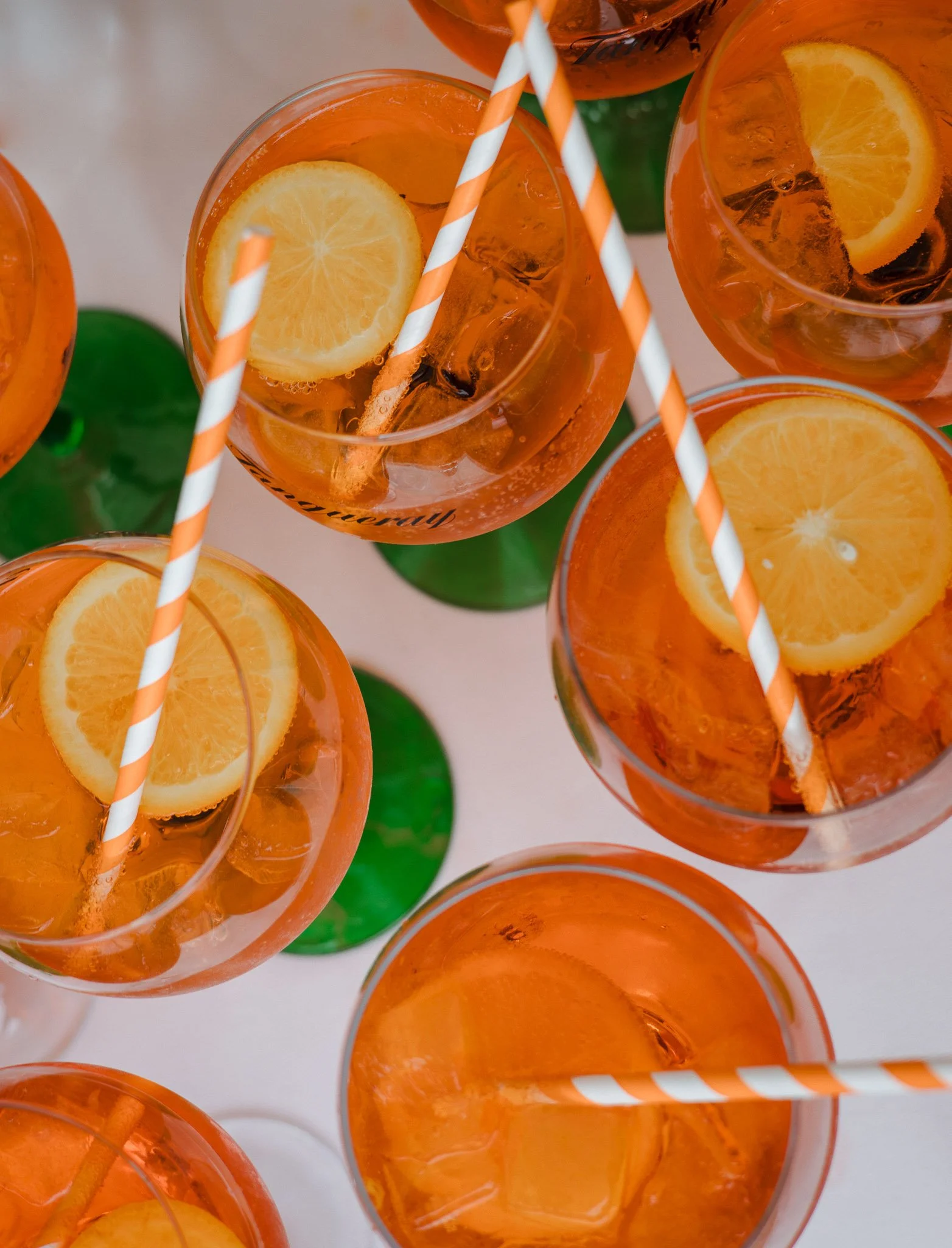 Top-down view of multiple glasses filled with orange-colored drinks, garnished with lemon slices and ice, with striped orange and white straws, and green bottle bases visible.