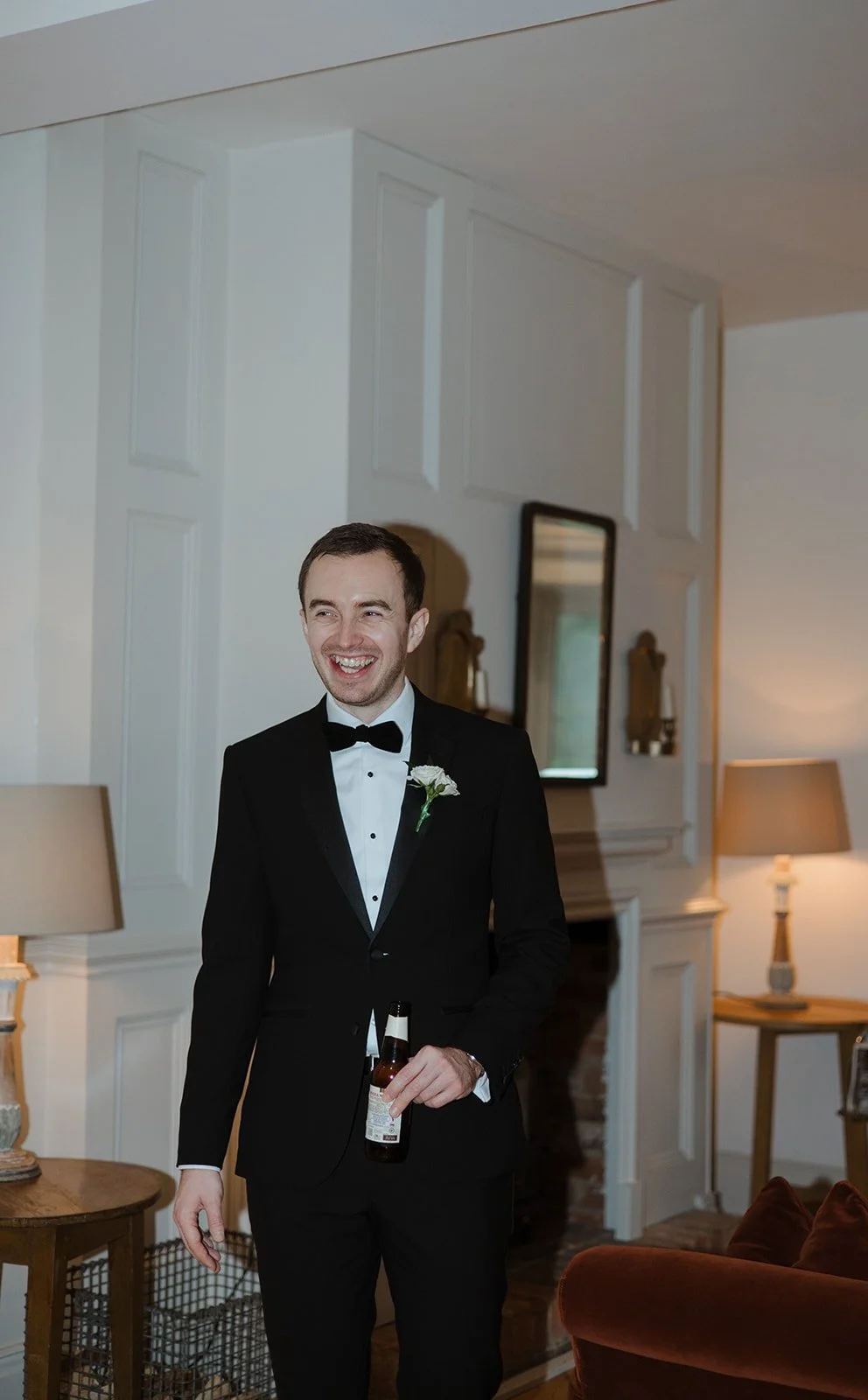 A man in a tuxedo with a boutonniere, holding a beer bottle, smiling in a warmly lit living room.
