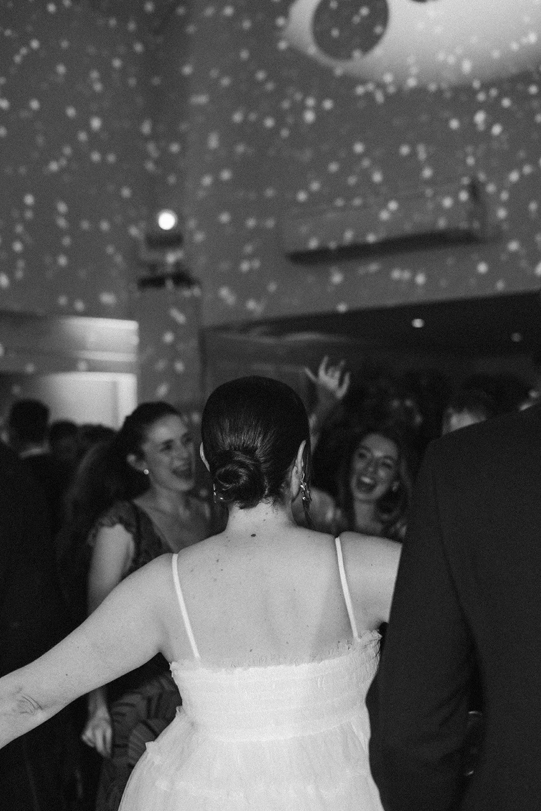 A woman with dark hair styled in a bun wearing a light-colored dress with spaghetti straps, dancing at a party. Two women in the background are smiling and enjoying the event, under a ceiling decorated with small star-like lights.
