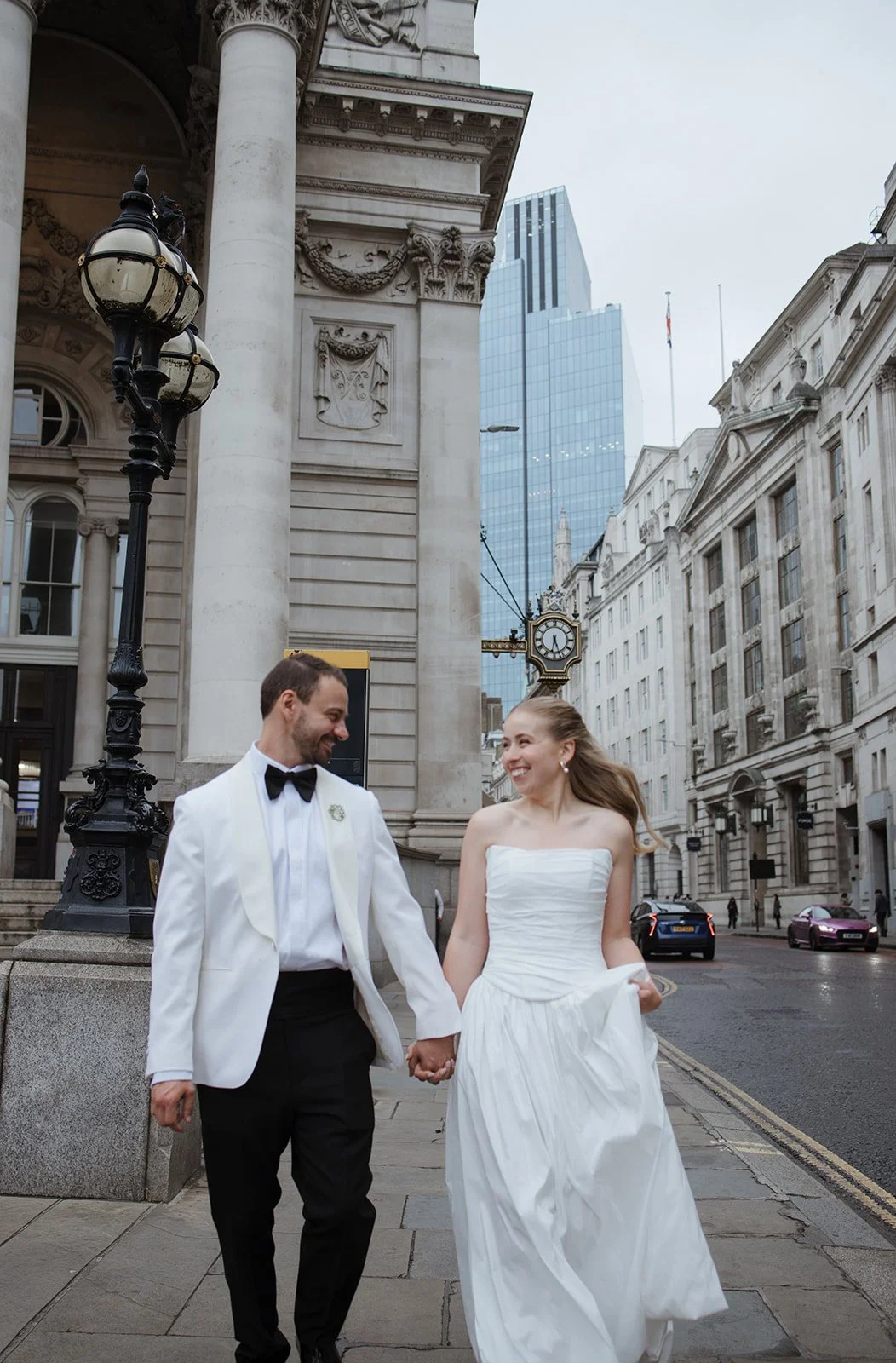 A newlywed couple walking hand in hand on a city street, smiling at each other. The groom is wearing a white tuxedo jacket with a black bow tie and the bride is in a strapless white wedding dress. Old stone buildings and modern glass skyscrapers are 