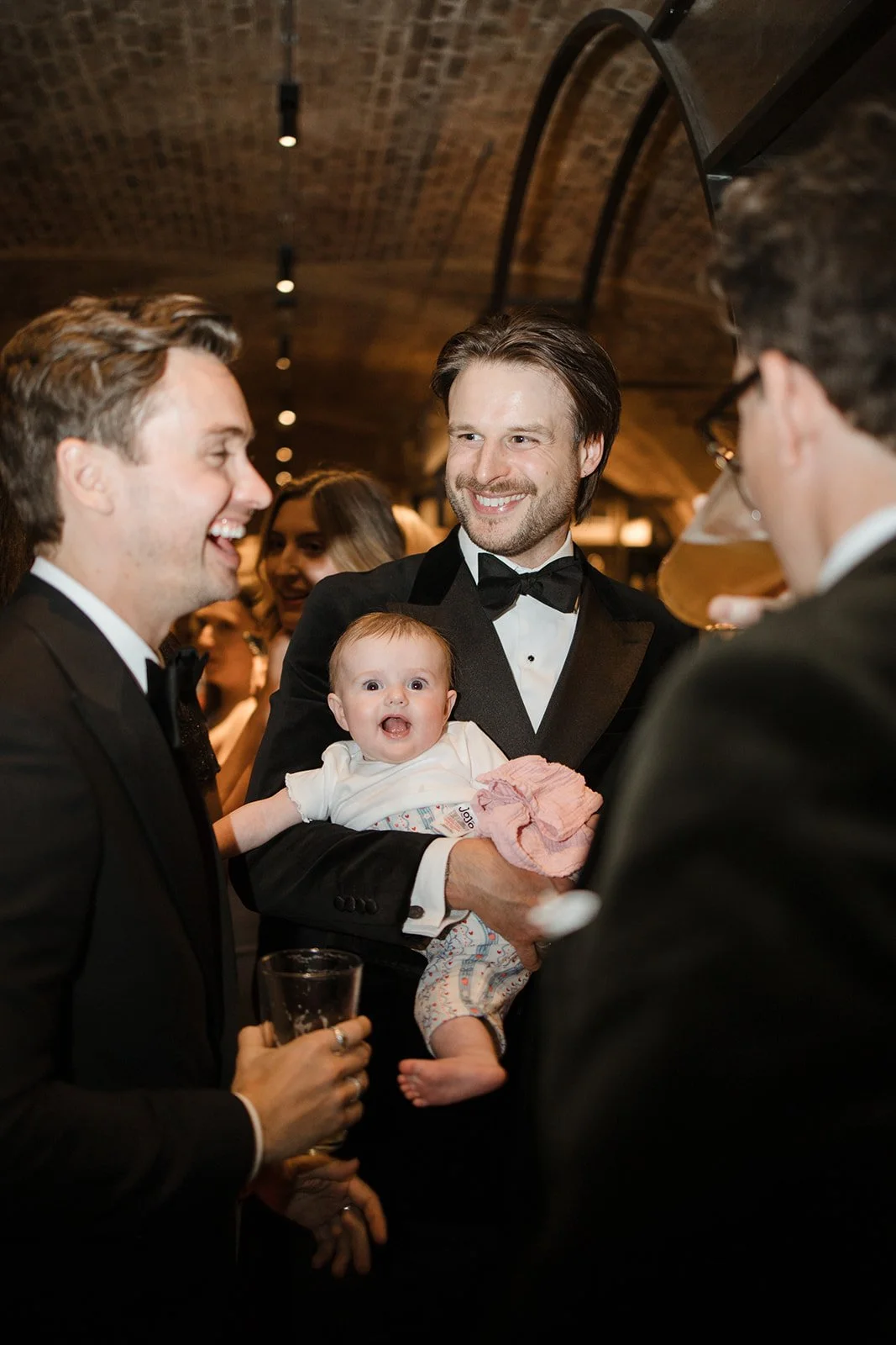 Group of people in tuxedos at a formal event, including a smiling man holding a surprised baby in a white outfit amid conversations, with a bar and warm lighting in the background.