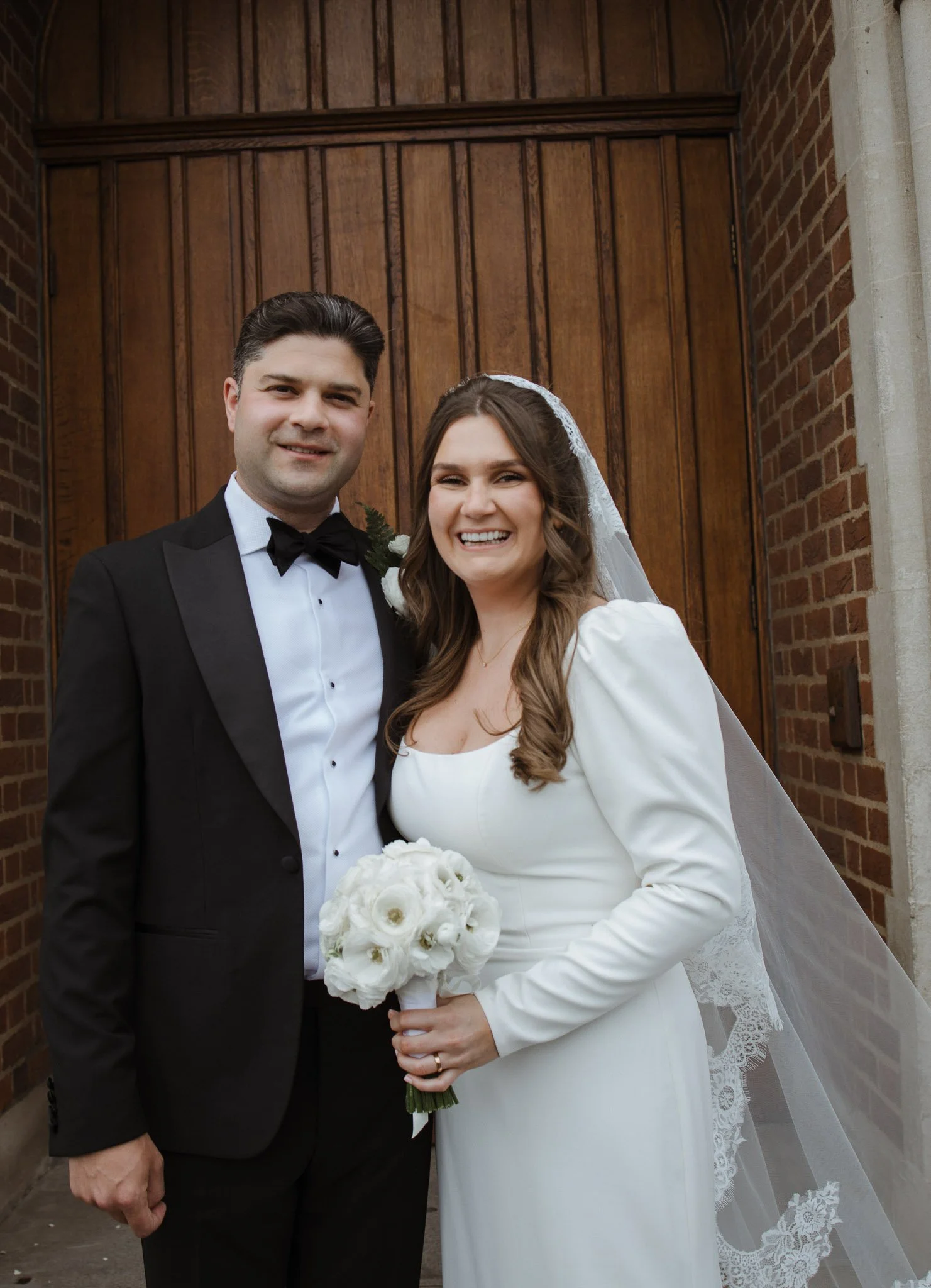 A bride and groom standing in front of a wooden door, smiling for a wedding photo. The bride holds a bouquet of white flowers and wears a white wedding dress with a veil. The groom wears a black tuxedo with a bow tie.