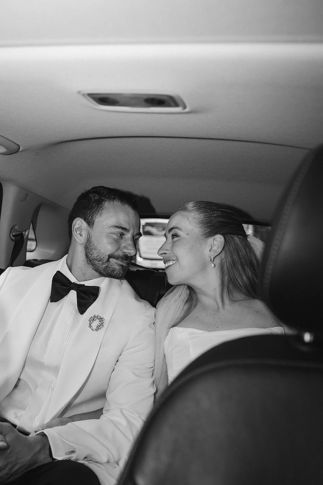 A black and white photo of a bride and groom sitting close together in the back of a limousine, smiling and looking at each other, dressed in wedding attire.
