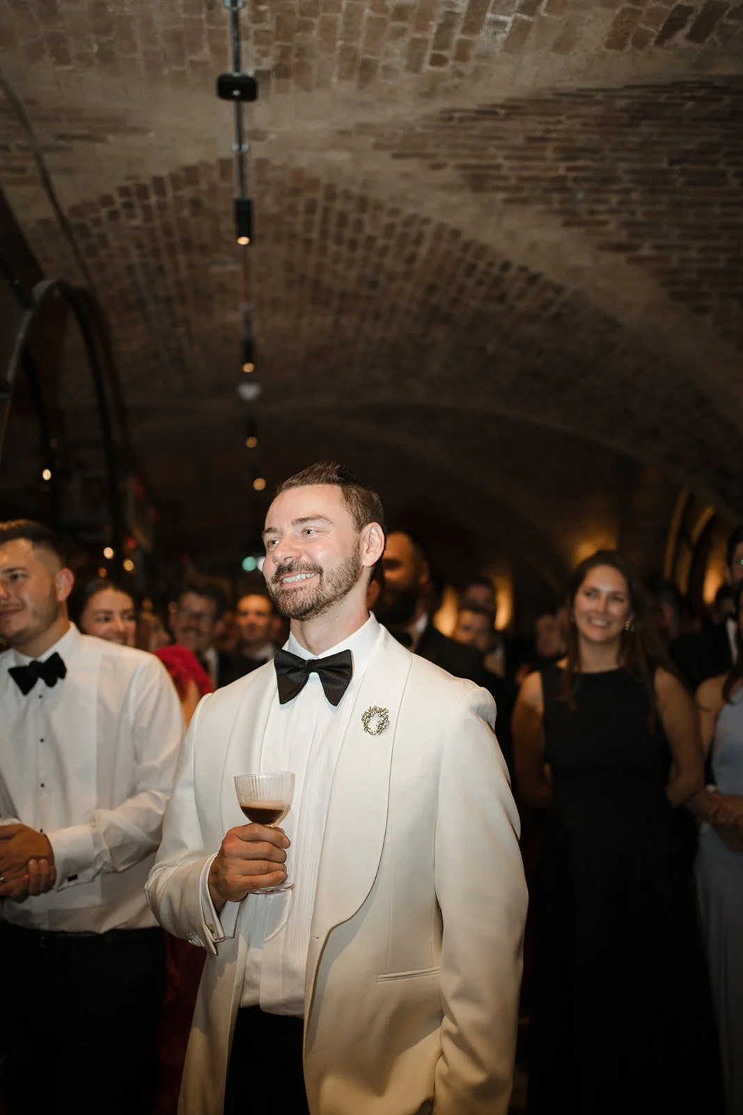 A man in a white tuxedo with a black bow tie and a floral lapel pin is holding a glass of red wine at a formal event, surrounded by other people dressed in formal attire in a dimly lit, brick-vaulted venue.