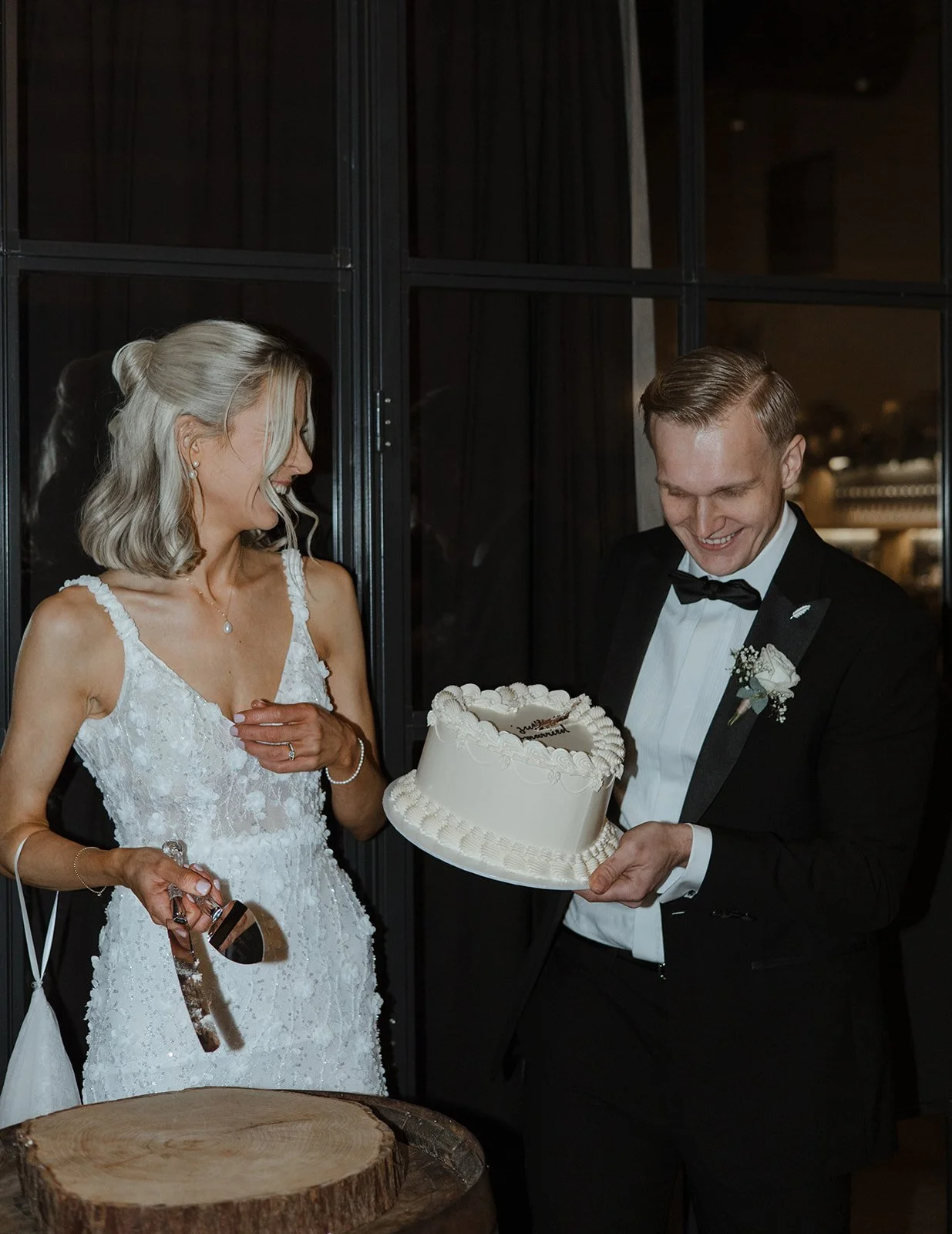 A bride and groom celebrating at a wedding, with the groom holding a cake and the bride with her hand on her chest, smiling.