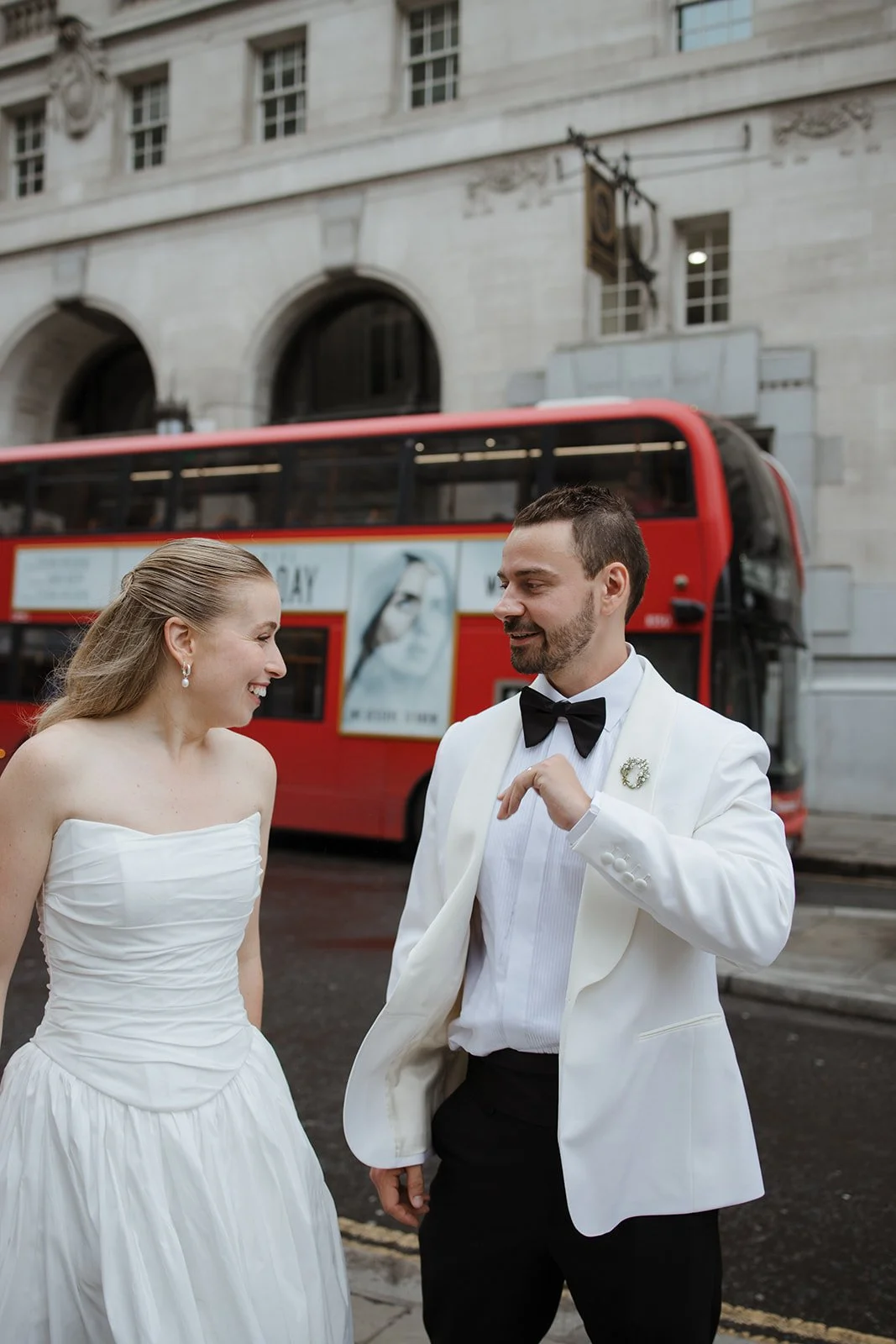 A bride and groom in wedding attire smiling and talking on a city street with a red double-decker bus in the background.
