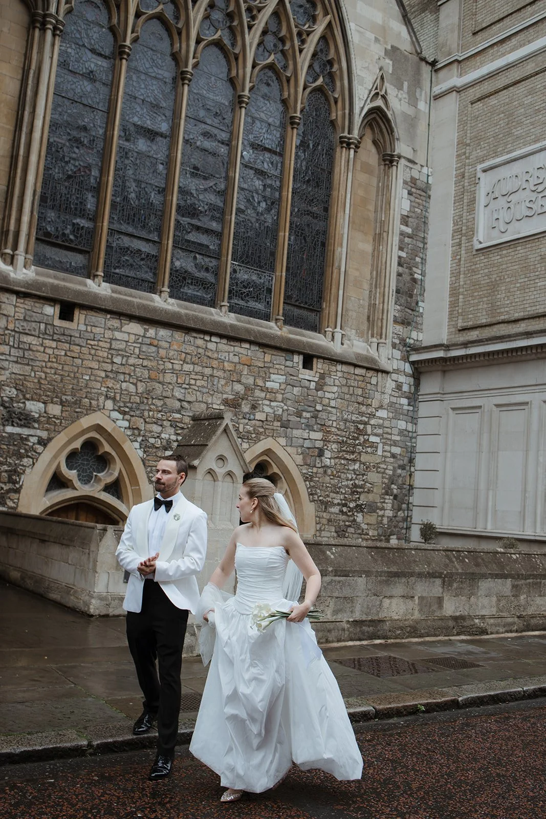 Bride and groom walking outside a stone church, with gothic windows, on a rainy day.