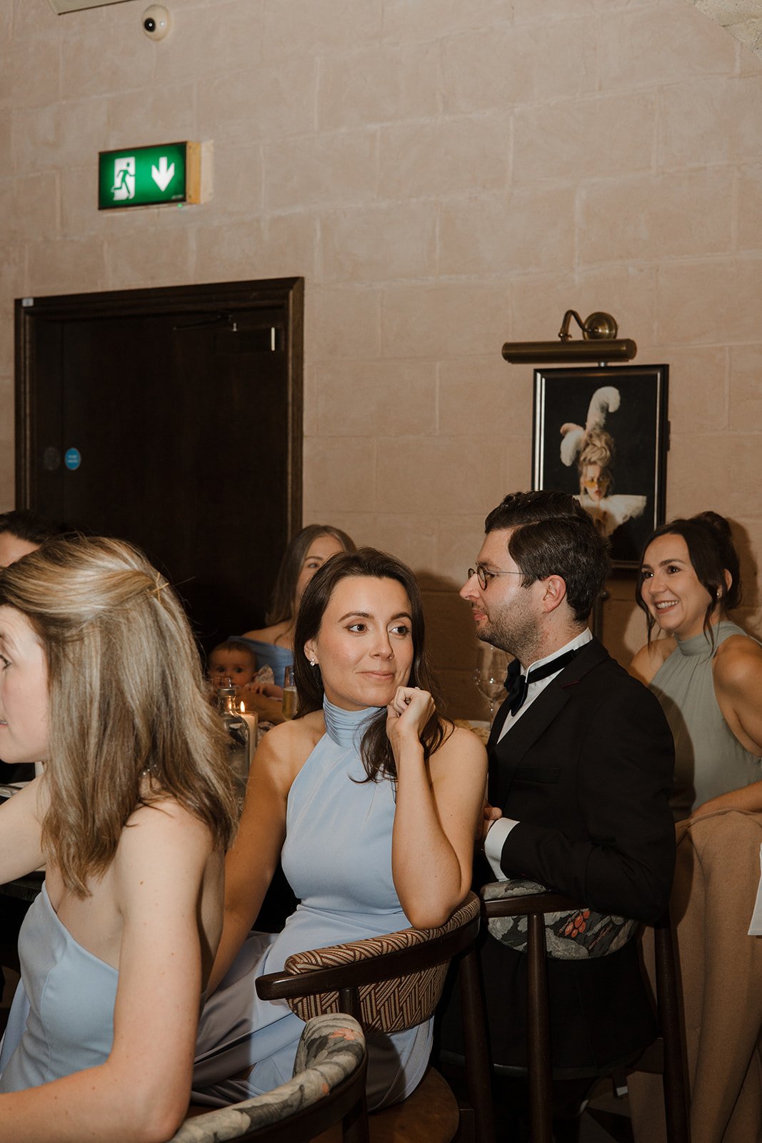 People at a formal gathering, including a woman in a gray dress sitting next to a man in a tuxedo, positioned at an event with a beige wall, a framed dog portrait, and a green exit sign.