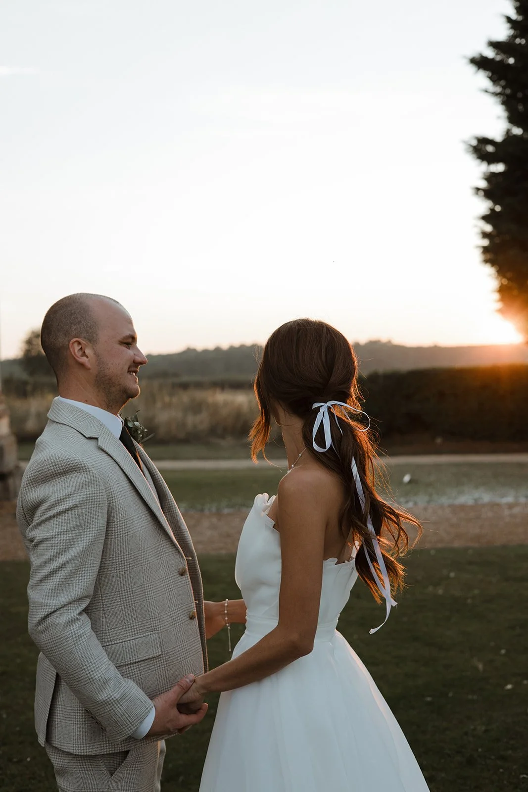 A bride and groom holding hands and facing each other outdoors at sunset, with a landscape of grass, trees, and rolling hills in the background.