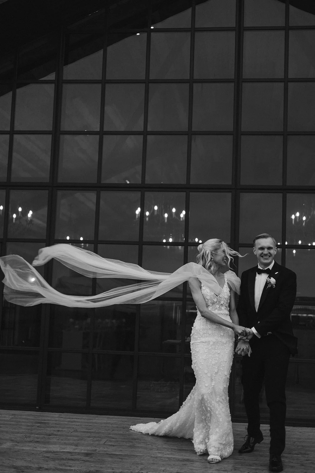 A couple on their wedding day holding hands and smiling, with the bride wearing a white lace gown and the groom in a black tuxedo, inside a building with a large glass window and chandeliers reflected in the glass.