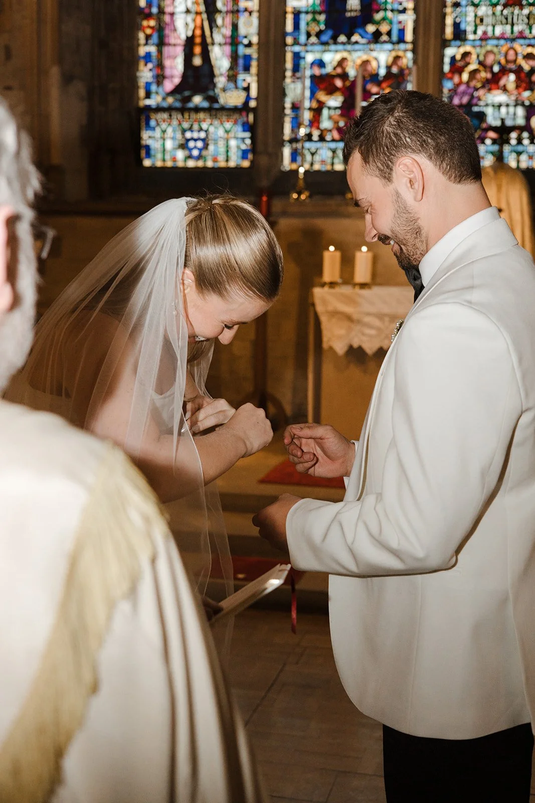A bride and groom exchanging rings during their wedding ceremony inside a church with stained glass windows.