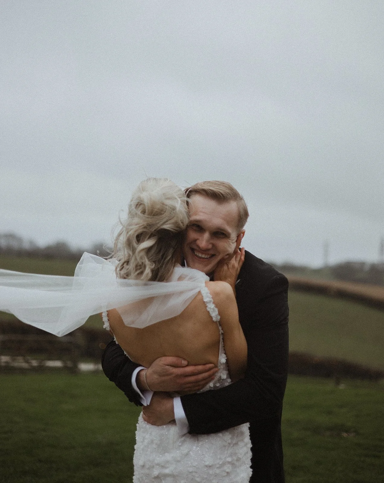 A couple on their wedding day hugging outdoors, the bride wearing a white wedding dress and veil, the groom in a black suit, smiling happily.