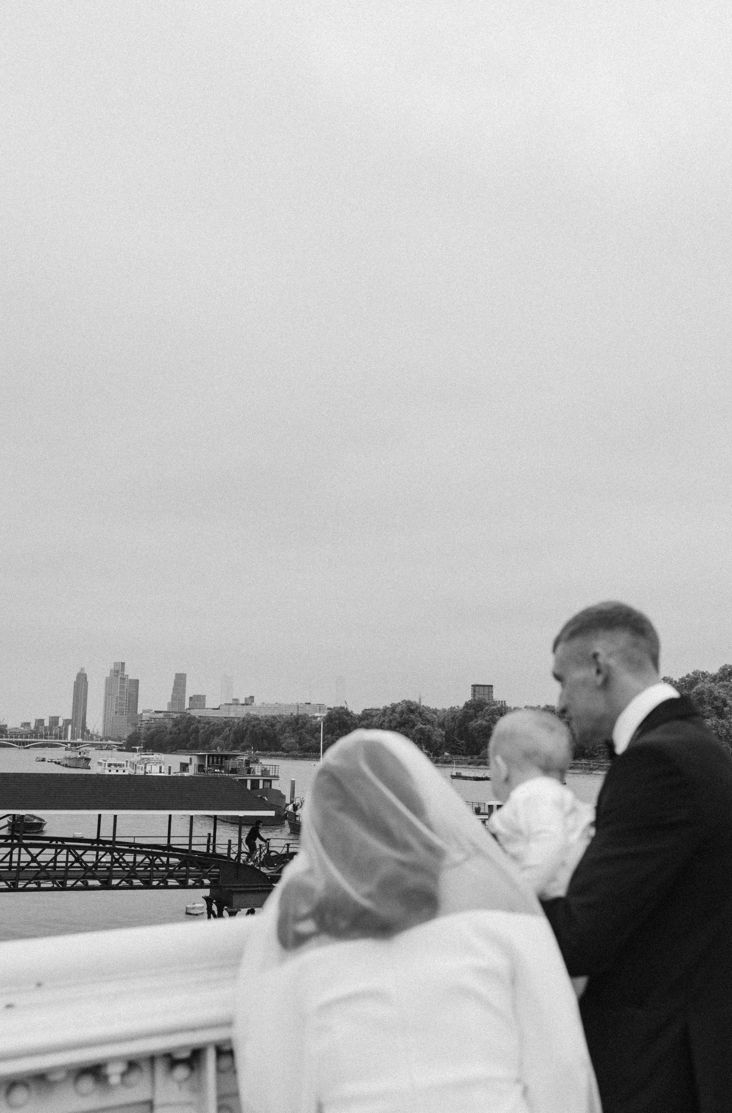 People dressed in formal attire, including women wearing headscarves and men in suits, gathered outdoors near a river, with a city skyline in the background.