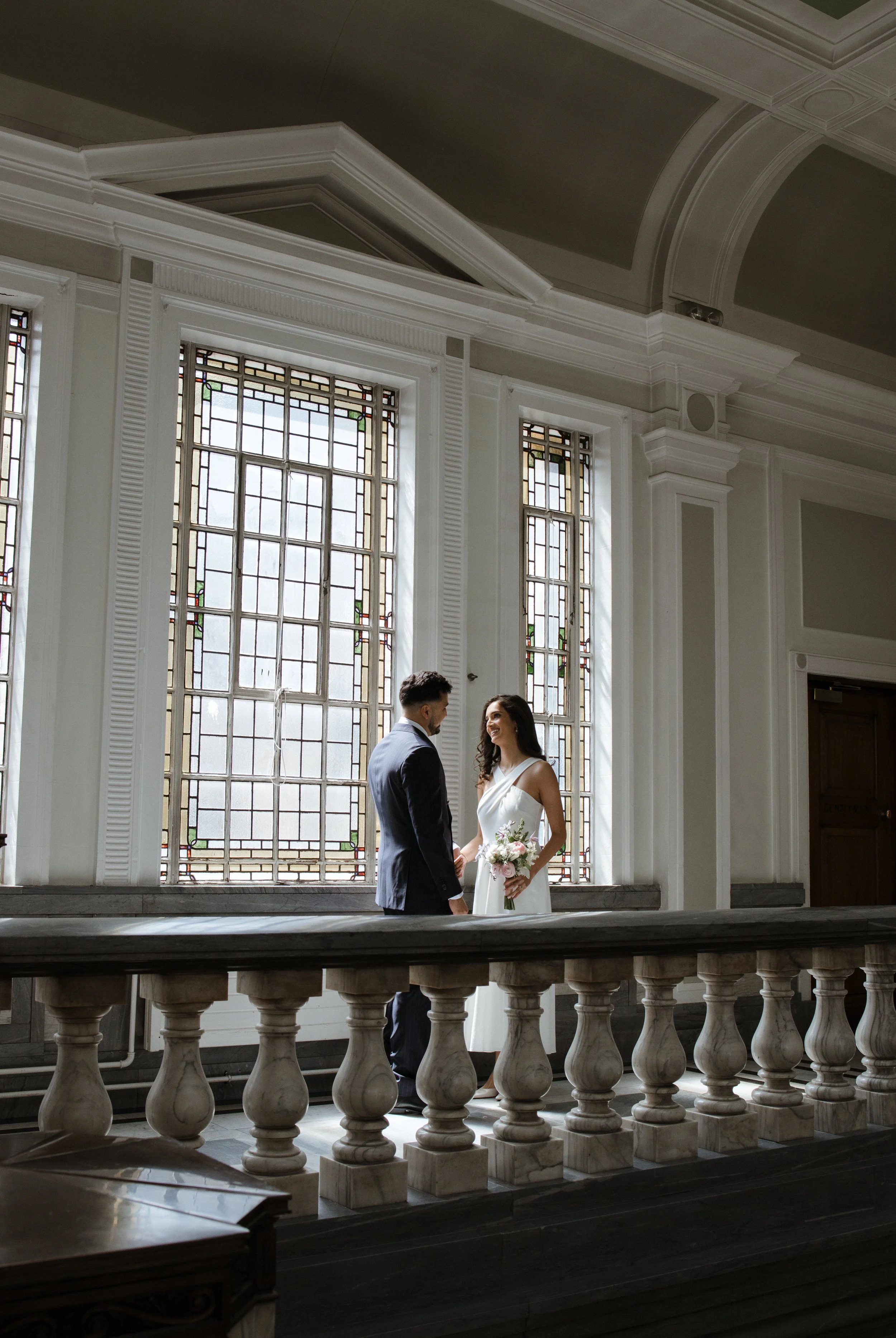 A bride and groom standing face to face, holding hands, inside a grand, historic building with large stained-glass windows, white walls, and decorative moldings.