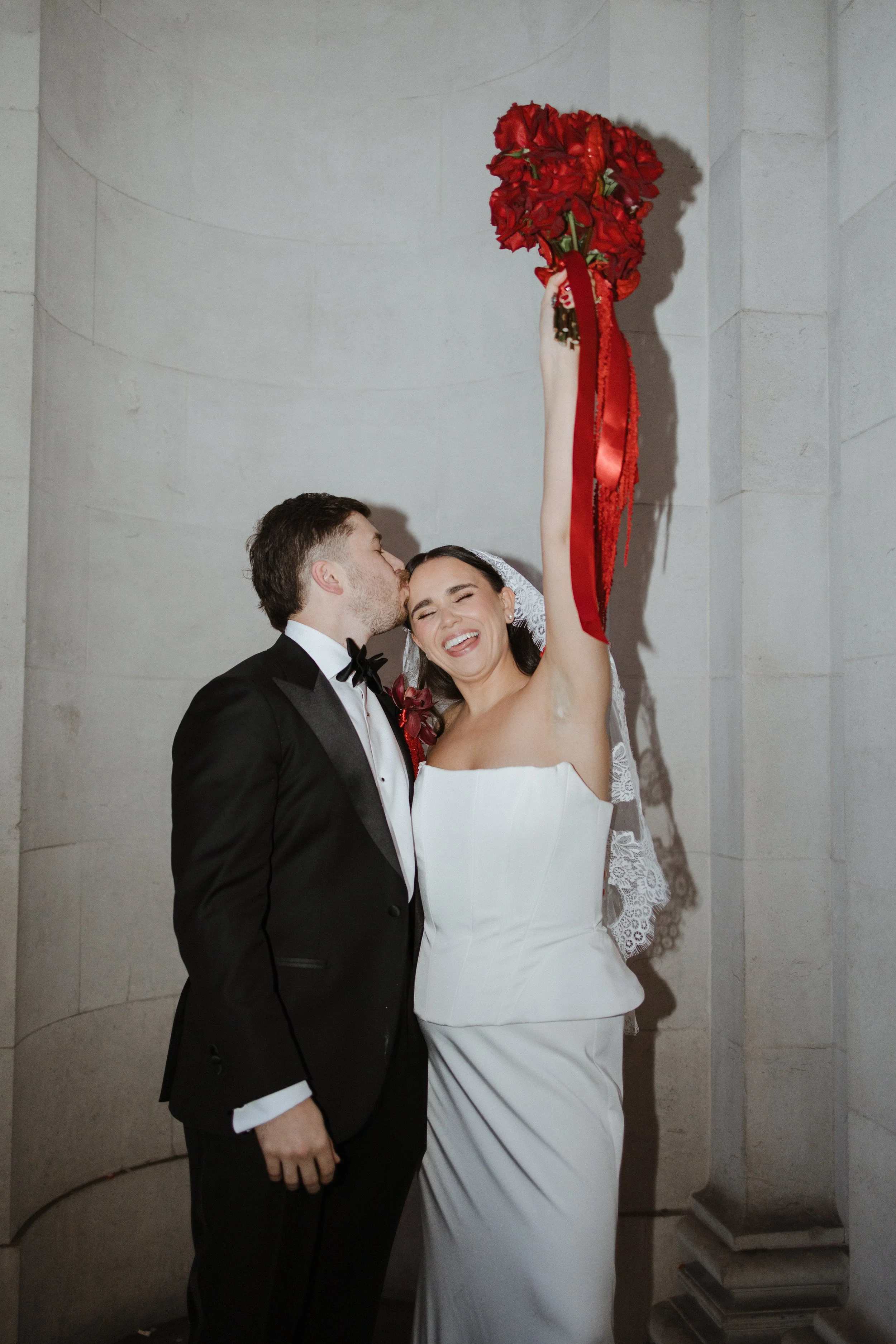 A bride and groom celebrating their wedding, with the bride holding a bouquet of red flowers high above her head while the groom kisses her forehead, both smiling affectionately.