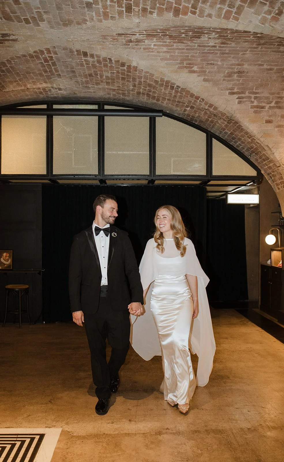 A bride and groom holding hands, walking together indoors with a brick ceiling, black curtains, and warm lighting.