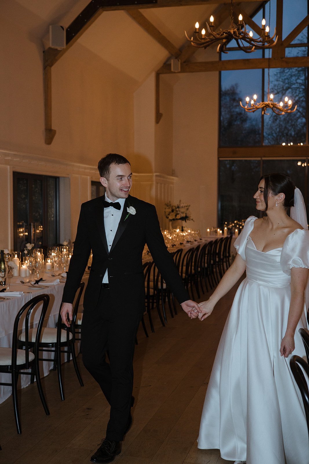 A bride and groom holding hands and dancing at their wedding reception in a decorated banquet hall with chandeliers, large windows, and candlelit tables.