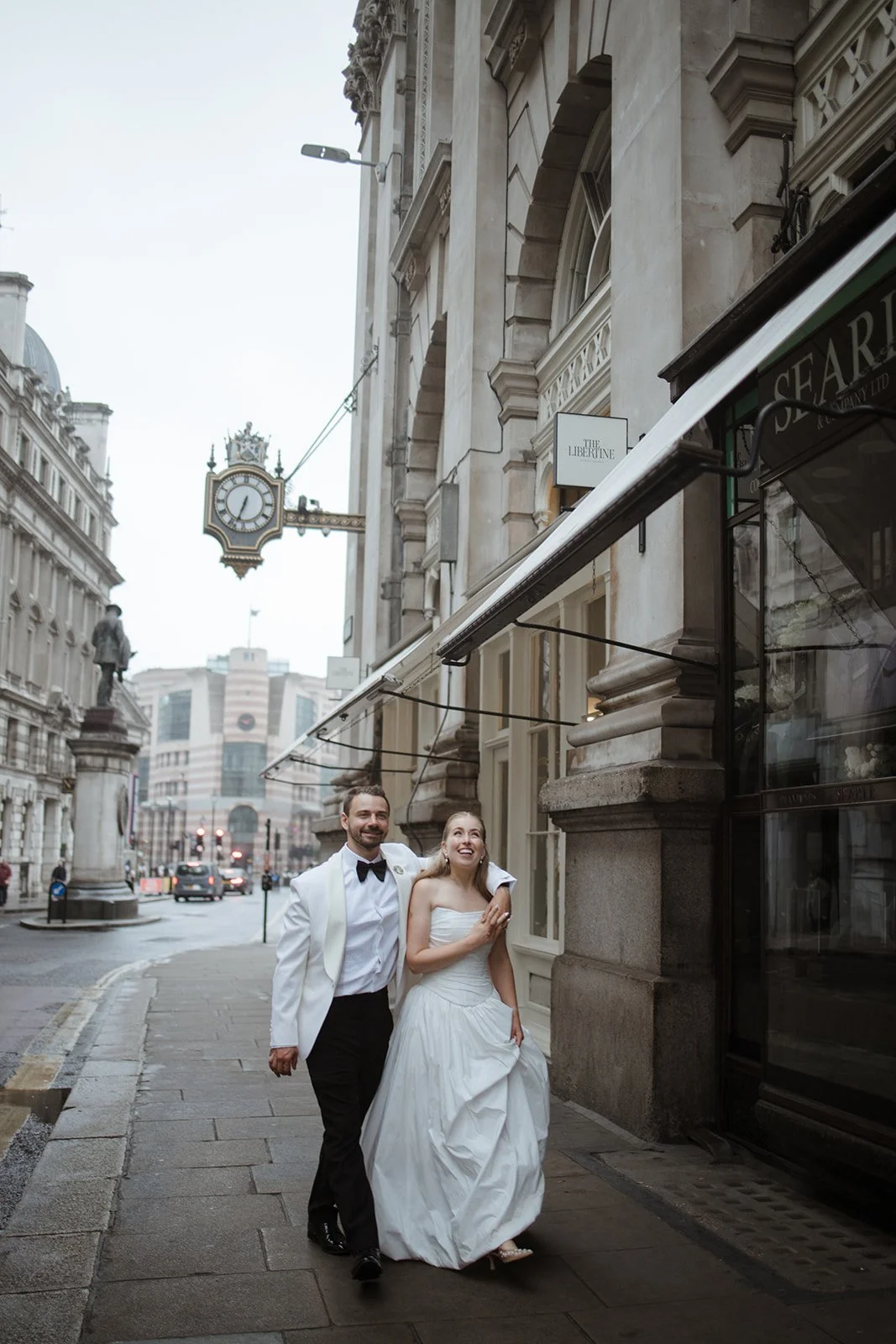 A smiling bride and groom in wedding attire walking on a city sidewalk with historic buildings, statues, and a clock in the background.