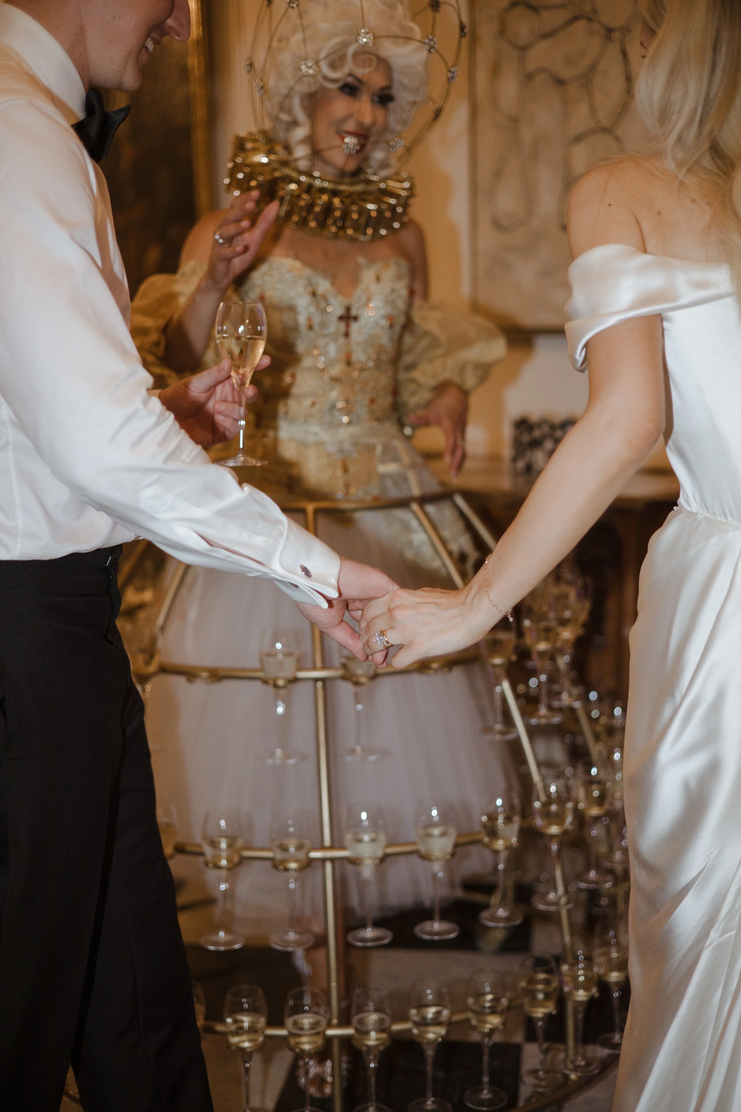 A couple holding hands during their wedding ceremony, with a woman in the background wearing an elegant dress and a decorative headpiece, holding a small mirror, in a warmly decorated room.