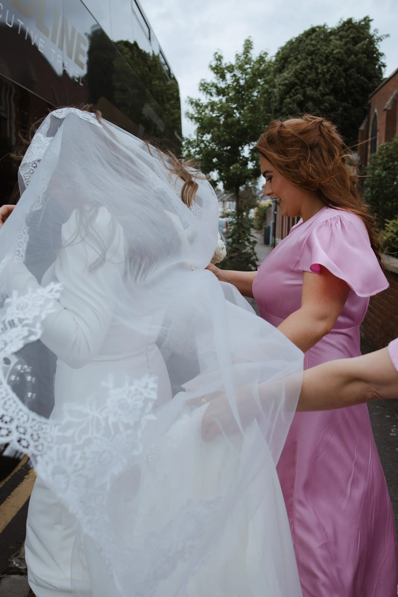 A woman in a pink dress helping another woman, dressed in a wedding gown, adjust her veil outdoors on a sidewalk, with trees and buildings in the background.