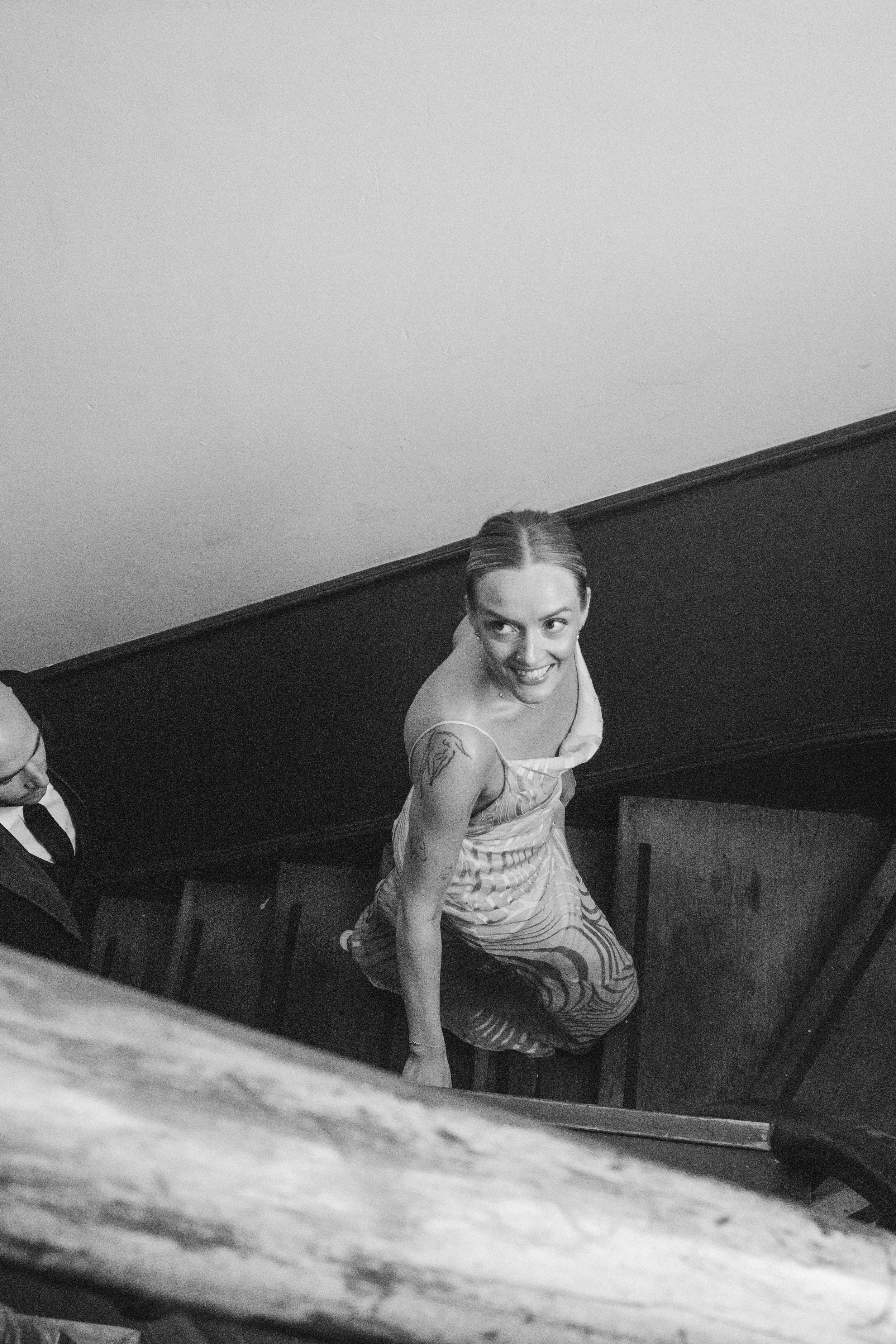 A woman in a, striped dress, is looking up and smiling while standing in a room with wooden furnishings and a plain wall. The image is black and white.