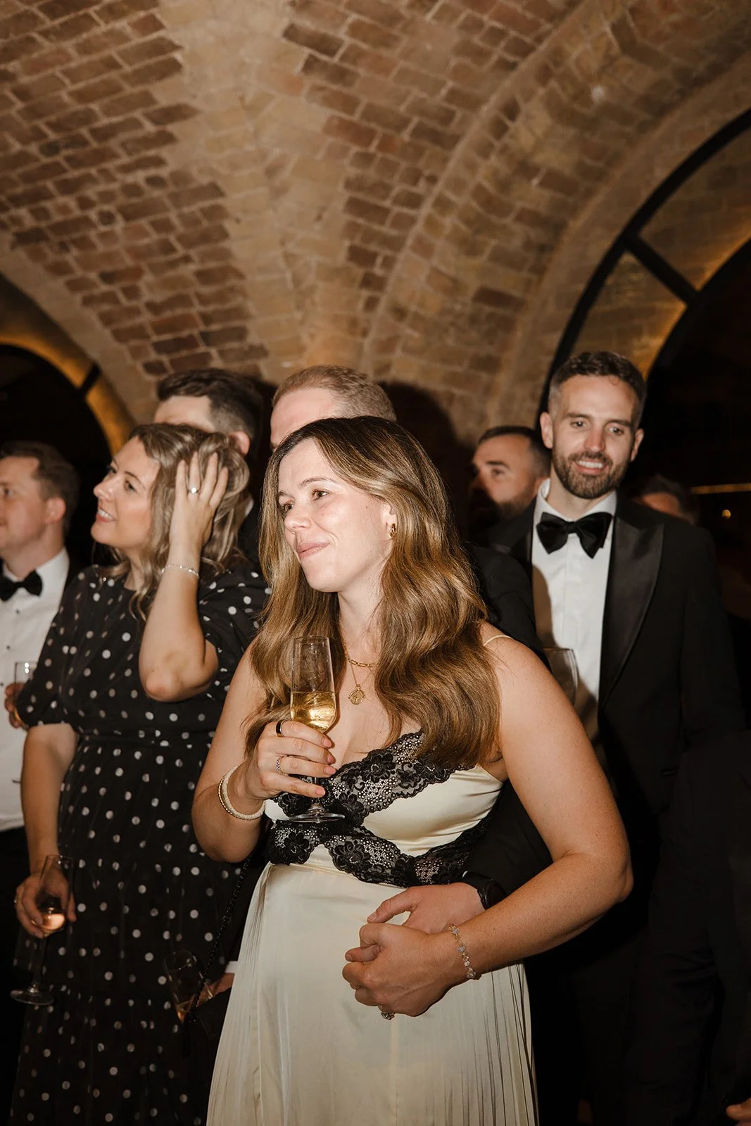 People dressed in formal attire, including a woman in a white dress with black lace, holding a glass of champagne at a social event in a venue with arched brick ceilings.