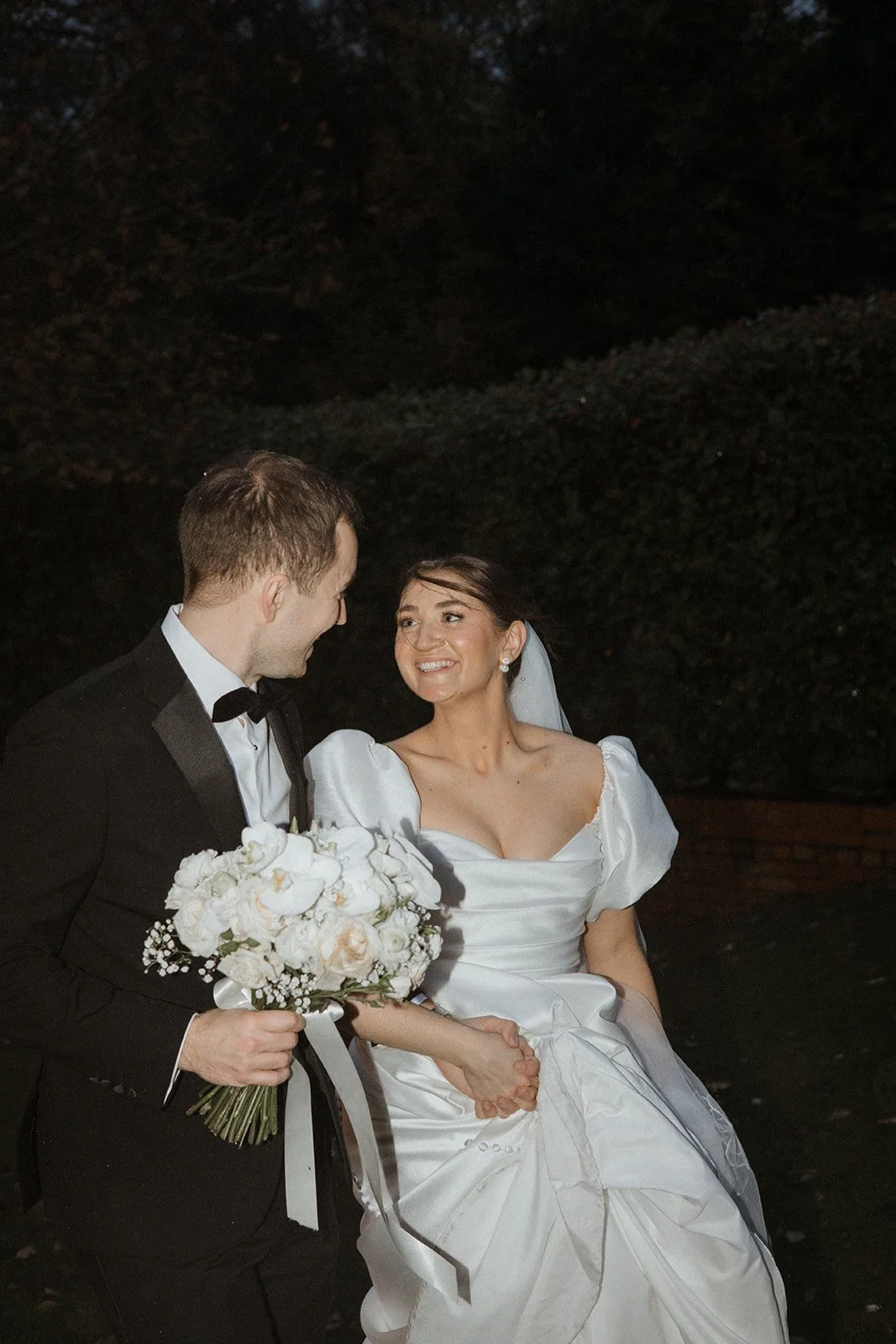 A bride in a white wedding gown holding hands with a groom in a black tuxedo, holding a bouquet of white flowers, smiling at each other during a wedding ceremony outdoors at night.