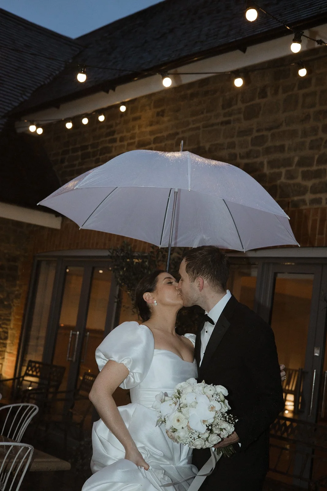 A bride and groom sharing a kiss under an umbrella during their wedding reception at night, with string lights overhead and a brick building in the background.