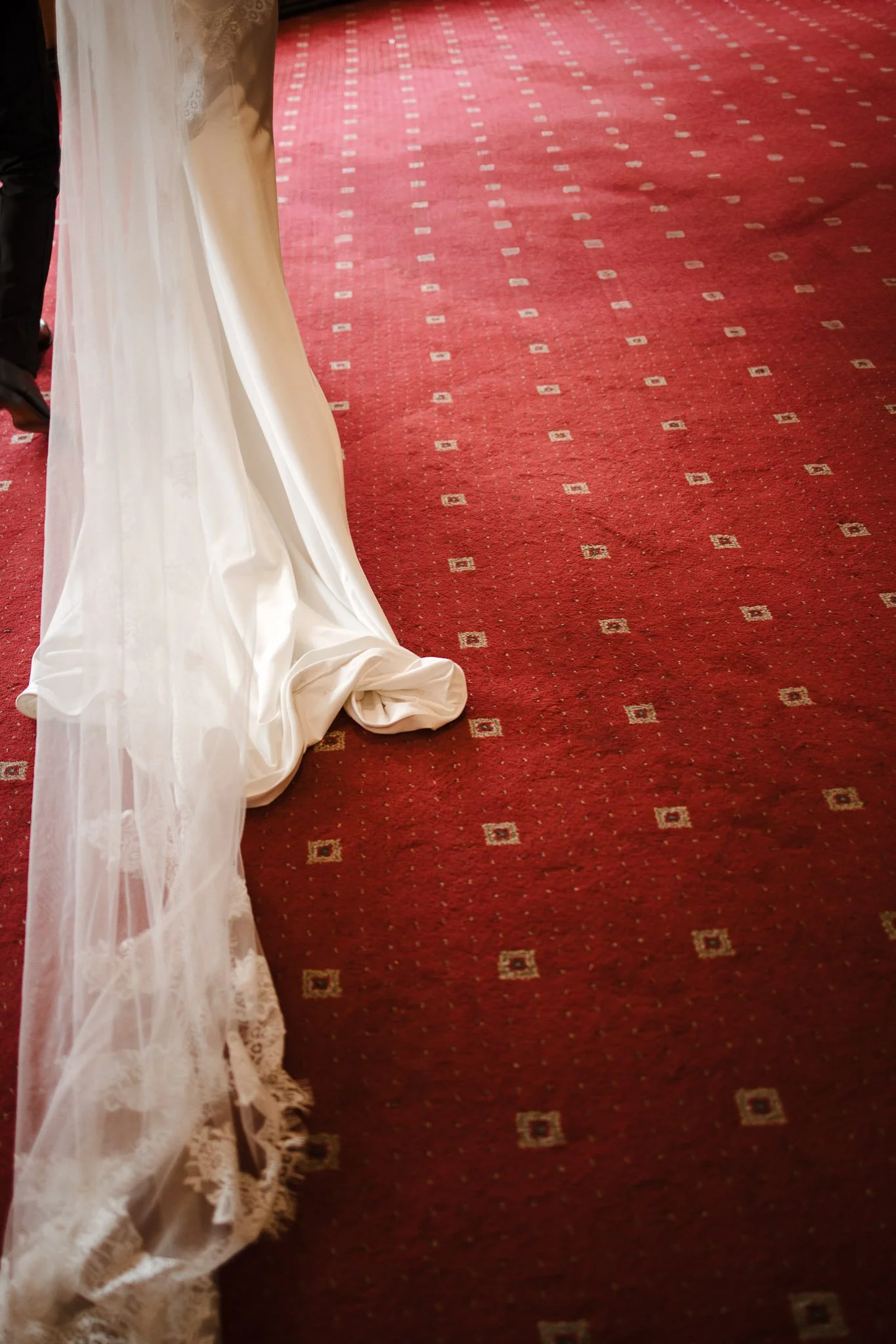 Part of a wedding dress with a long train on a patterned red carpet.