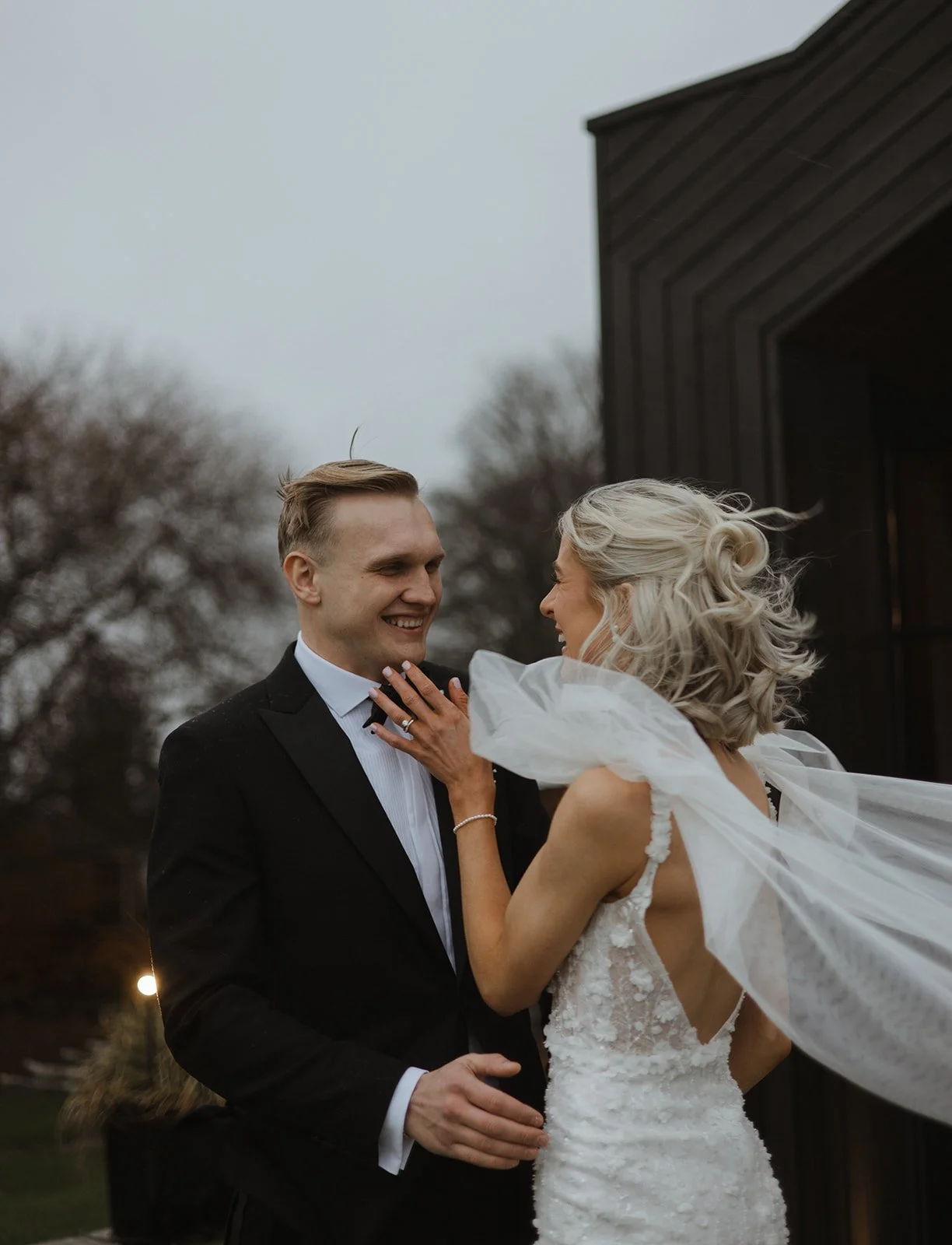 A bride and groom smiling and embracing outdoors on a cloudy day, with trees in the background.