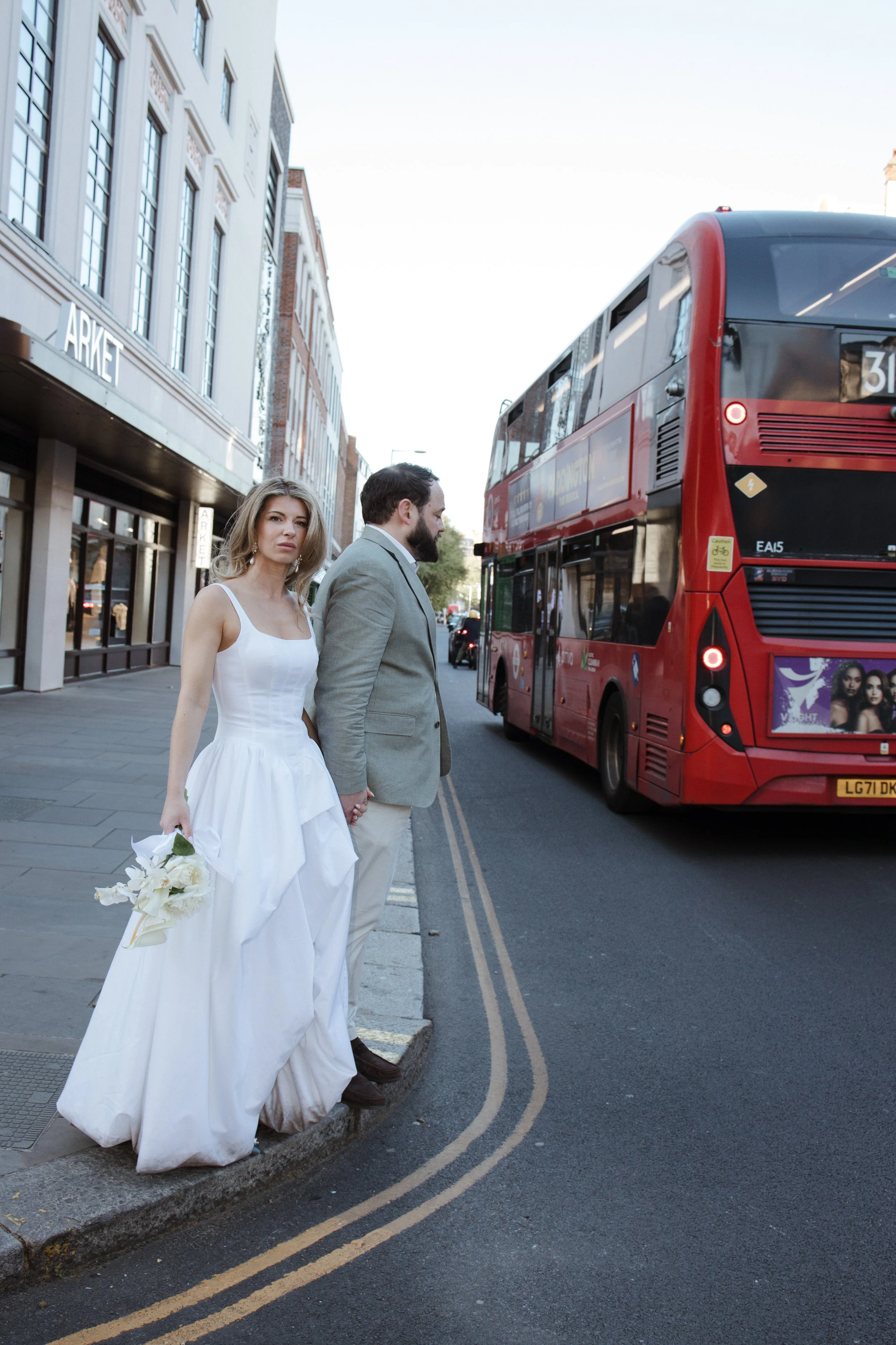 A bride in a white wedding dress and a groom in a beige suit holding hands on a city street, standing beside a red double-decker bus, with buildings and shopfronts in the background.