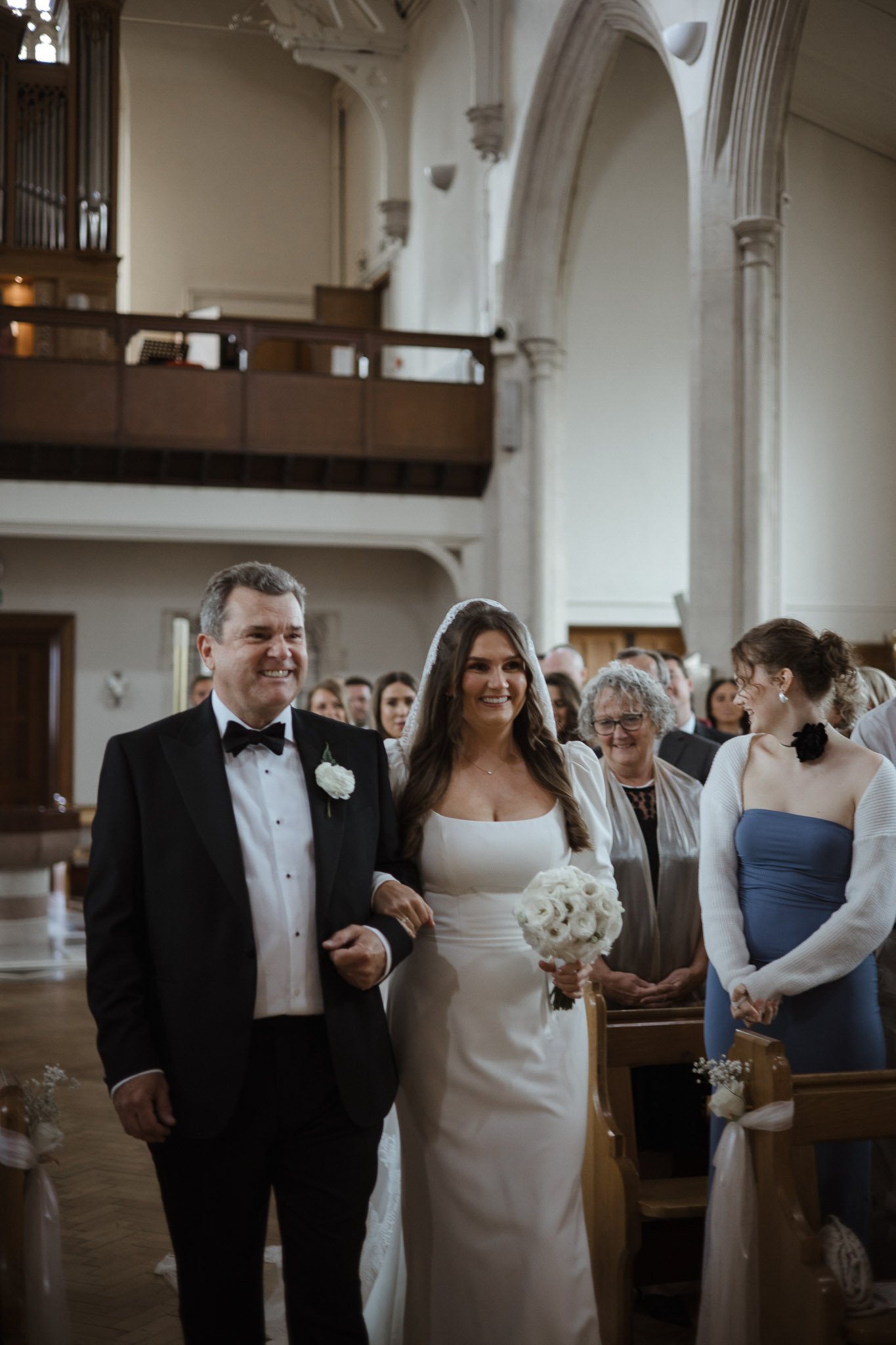 A bride walking down the aisle with her father at a wedding ceremony inside a church, surrounded by guests.