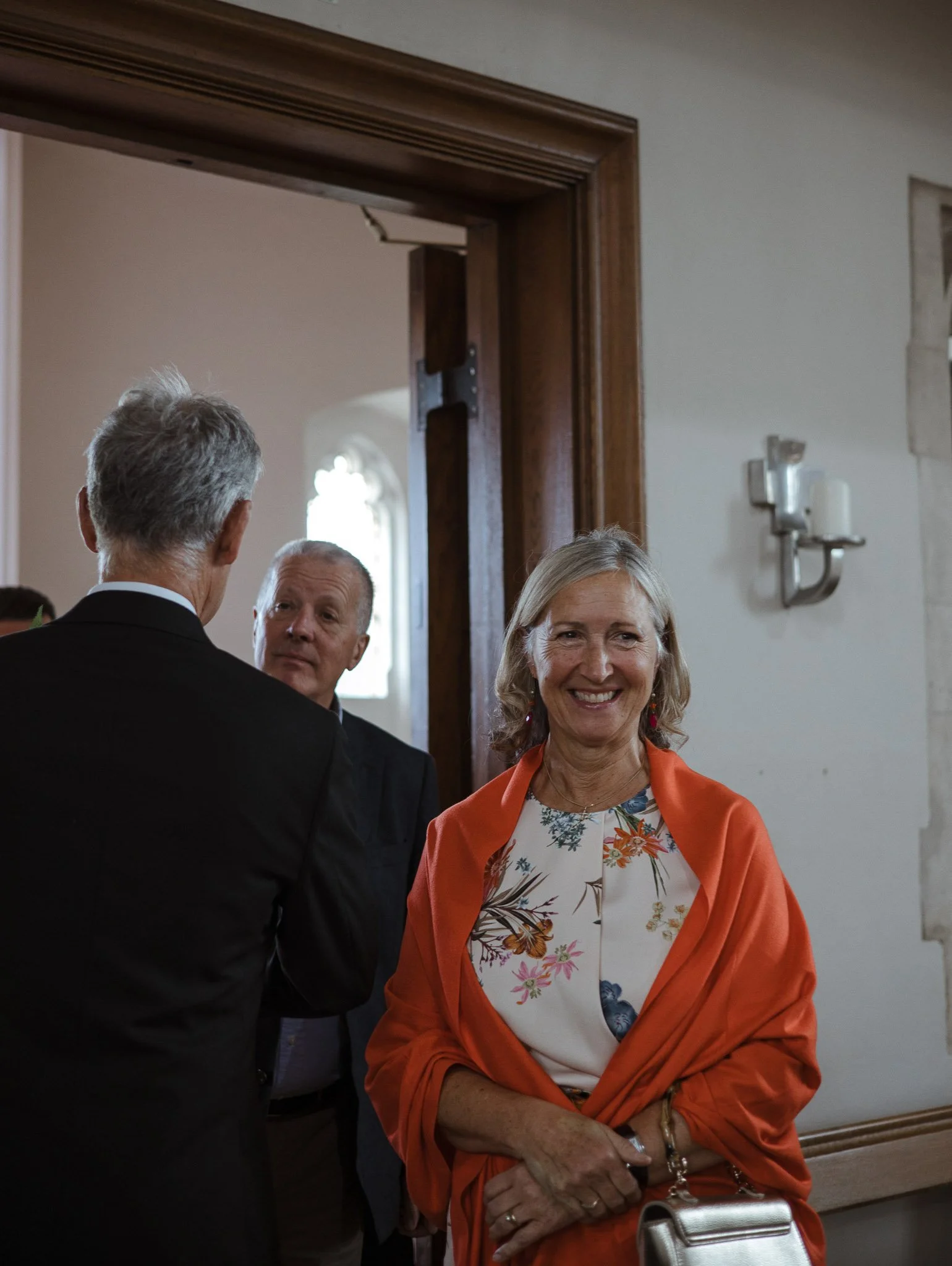 A woman smiling warmly while dressed in a floral top and orange shawl, standing indoors at a social gathering, with two men in suits conversing behind her.