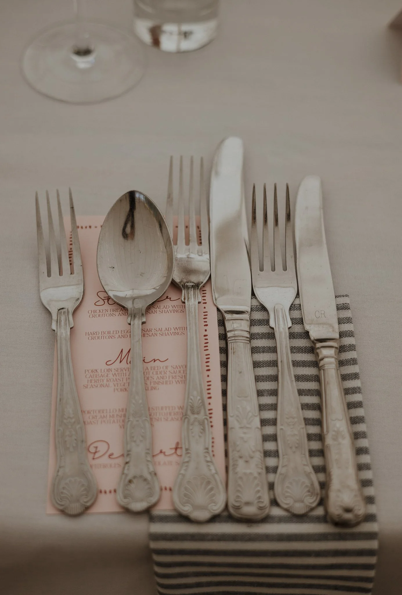 Set of six silverware pieces including three forks, one spoon, and two knives, arranged on a striped napkin on a table with a menu underneath, with a wine glass and water glass in the background.