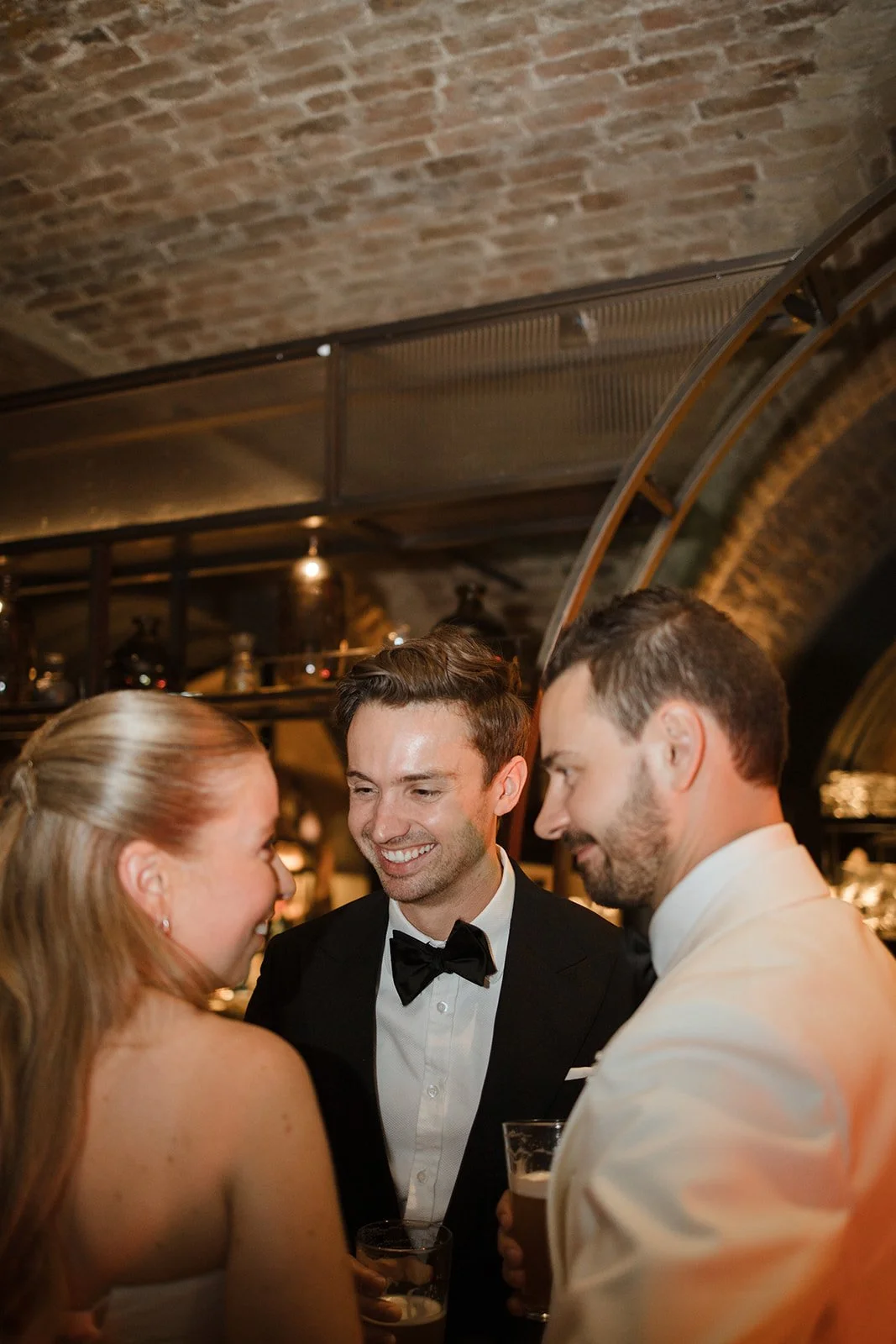 Three people dressed in formal attire laughing and talking at a social event.