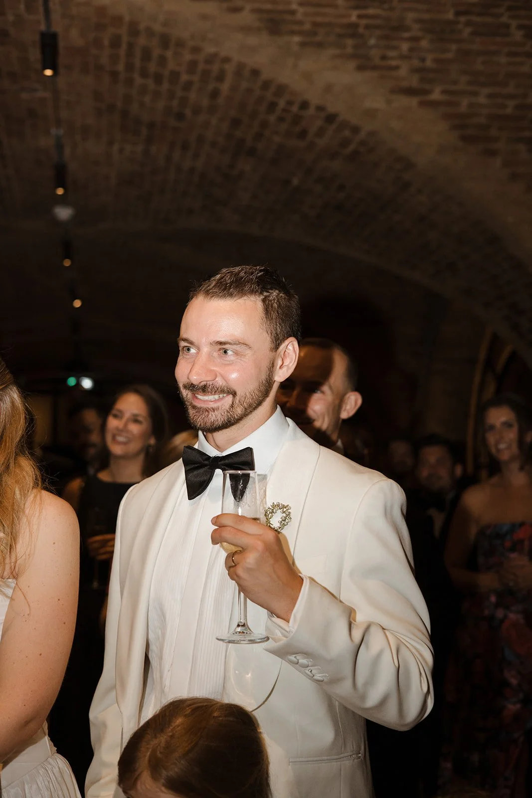 A man in a white tuxedo with a black bow tie holding a champagne flute at a formal event, smiling among other smiling guests in the background.