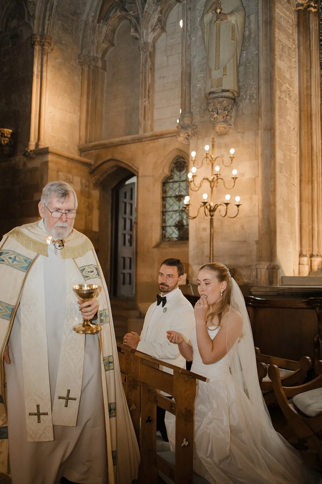 A wedding ceremony inside a church with a priest, a groom, and a bride. The priest is holding a chalice, and the bride is wearing a wedding dress with a veil while the groom is dressed in a tuxedo. The church features stone walls, arched windows, a c