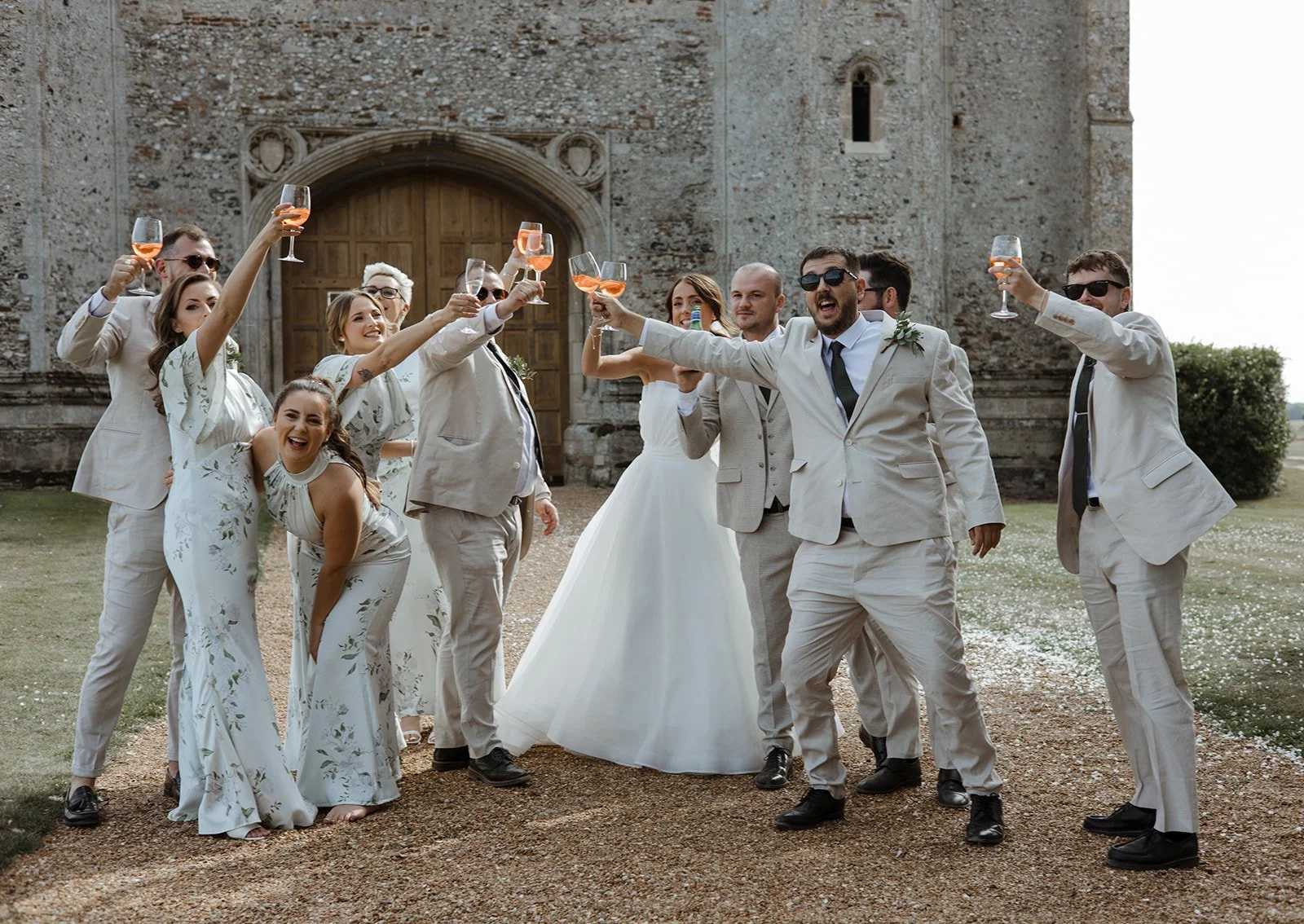 A group of people, including men in suits and women in white dresses, celebrating with glasses of rosé wine outside a historic stone building, likely at a wedding. They are smiling and raising their glasses in a toast.