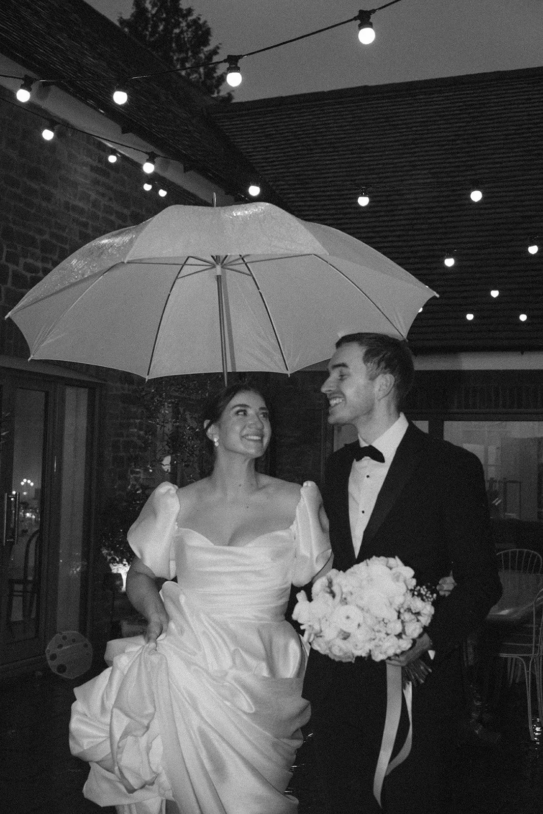 A black-and-white photo of a bride and groom standing close together under an umbrella during a rainy evening. The bride is in a wedding dress, holding the edge with one hand, and the groom is in a tuxedo holding a bouquet of flowers. Christmas or de