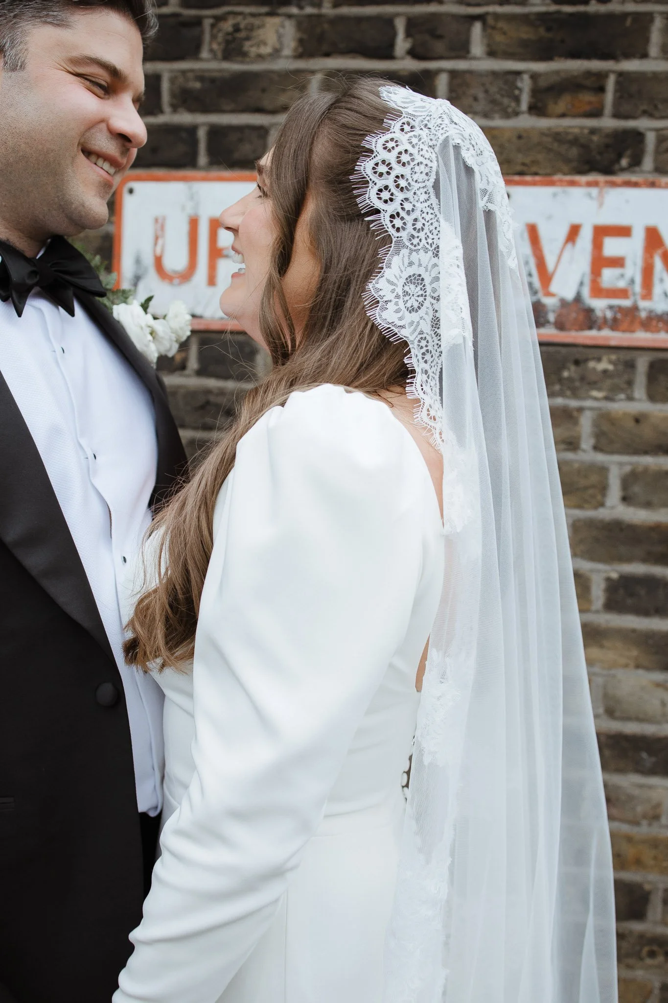 A bride and groom smiling at each other during their wedding, standing in front of a brick wall with a partially visible sign.