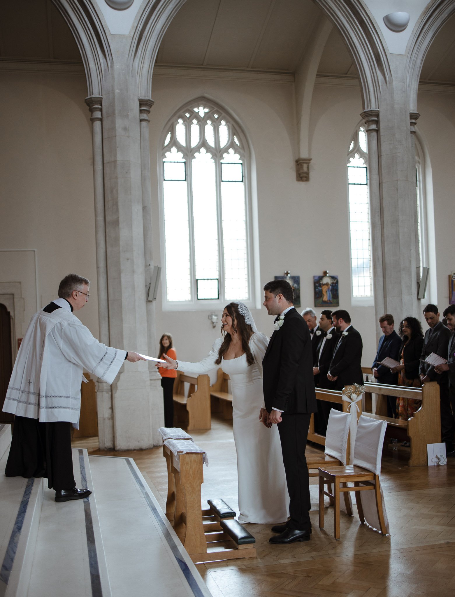 Bride and groom exchanging vows during wedding ceremony in church, officiant handing them a document.