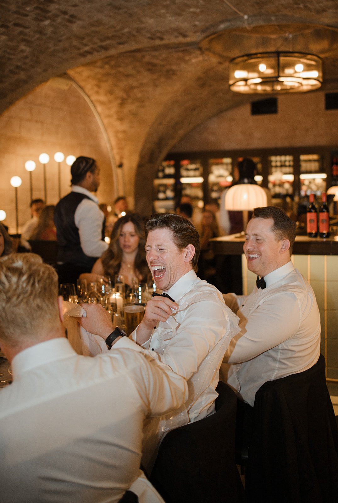People in formal attire cheering and enjoying drinks at a dinner event in an elegant restaurant with arched brick ceilings and warm lighting.