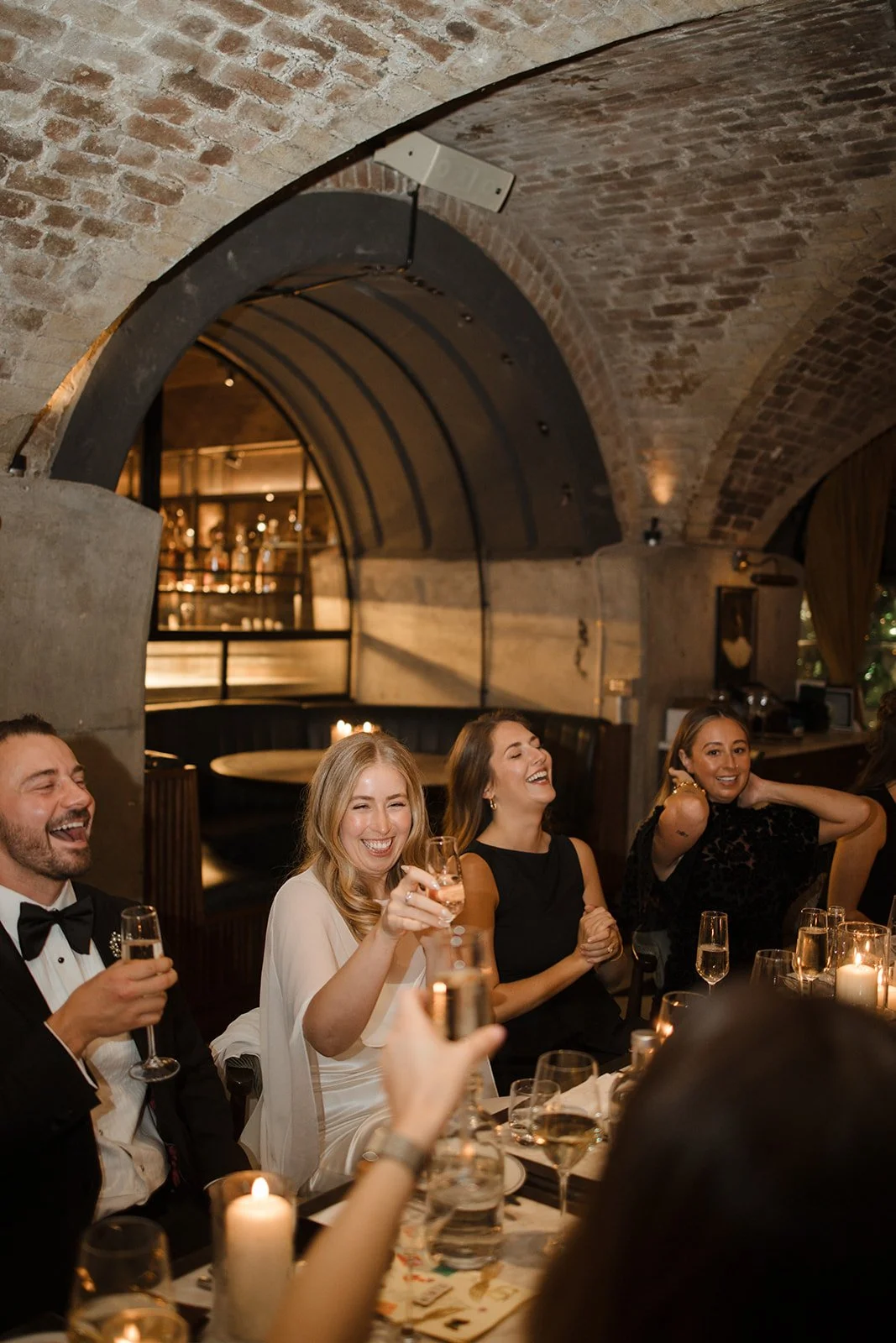 Group of four adults sitting at a dinner table in a dimly lit restaurant, smiling and laughing, holding glasses of champagne.