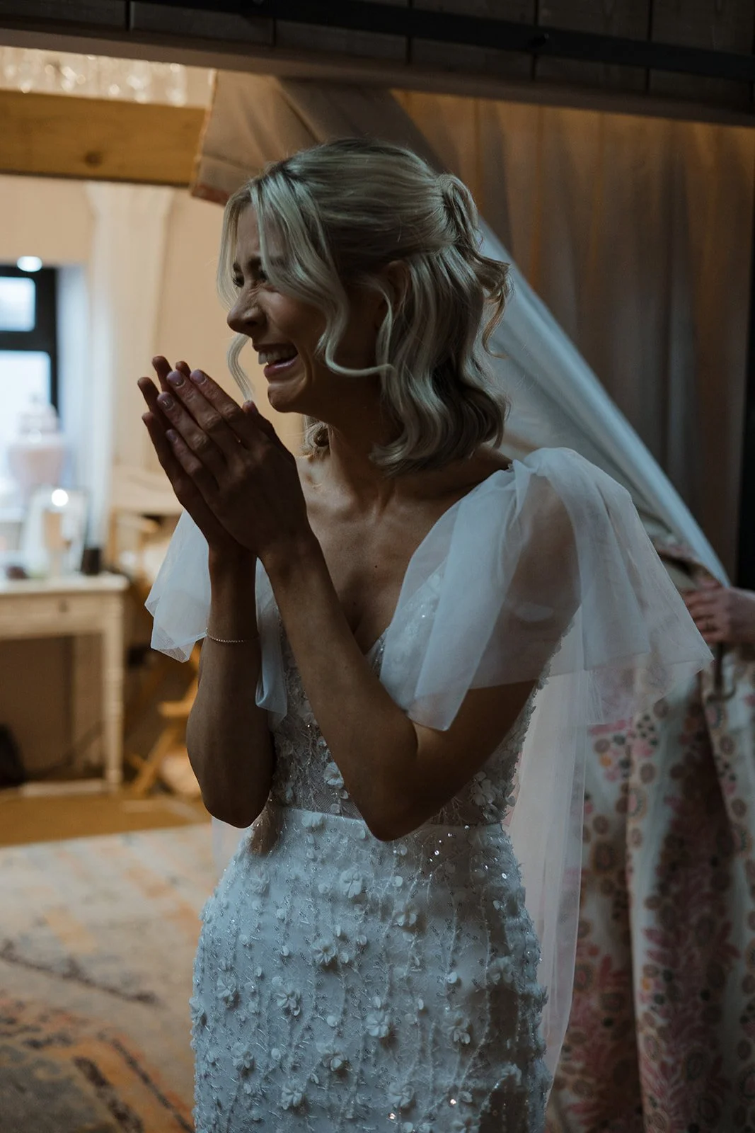A woman in a wedding dress is visibly emotional, clasping her hands near her face and smiling, likely at her wedding or a special moment.
