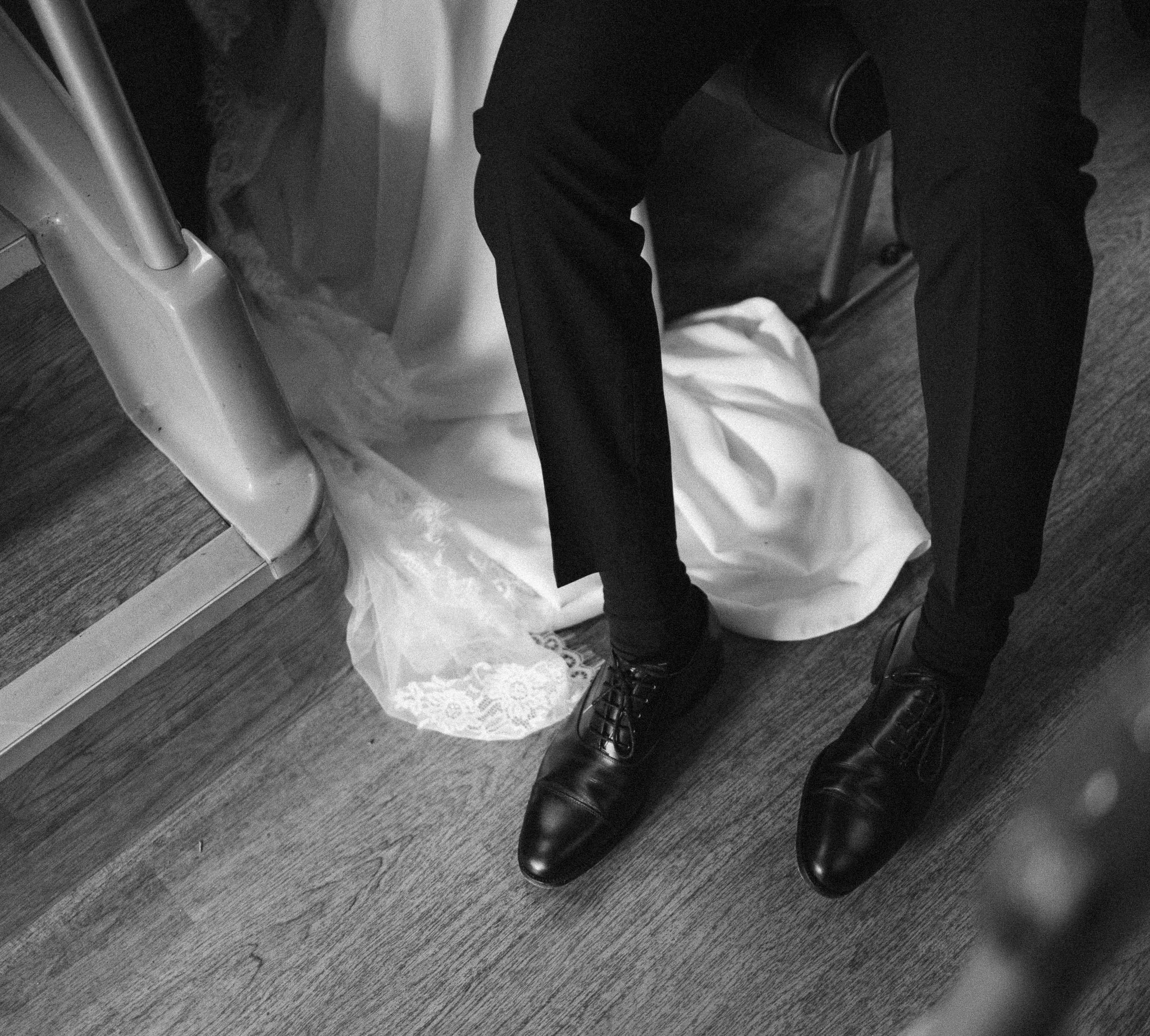 Black and white photo of a person in formal attire, sitting with legs apart, with a white lace cloth or veil on the floor.