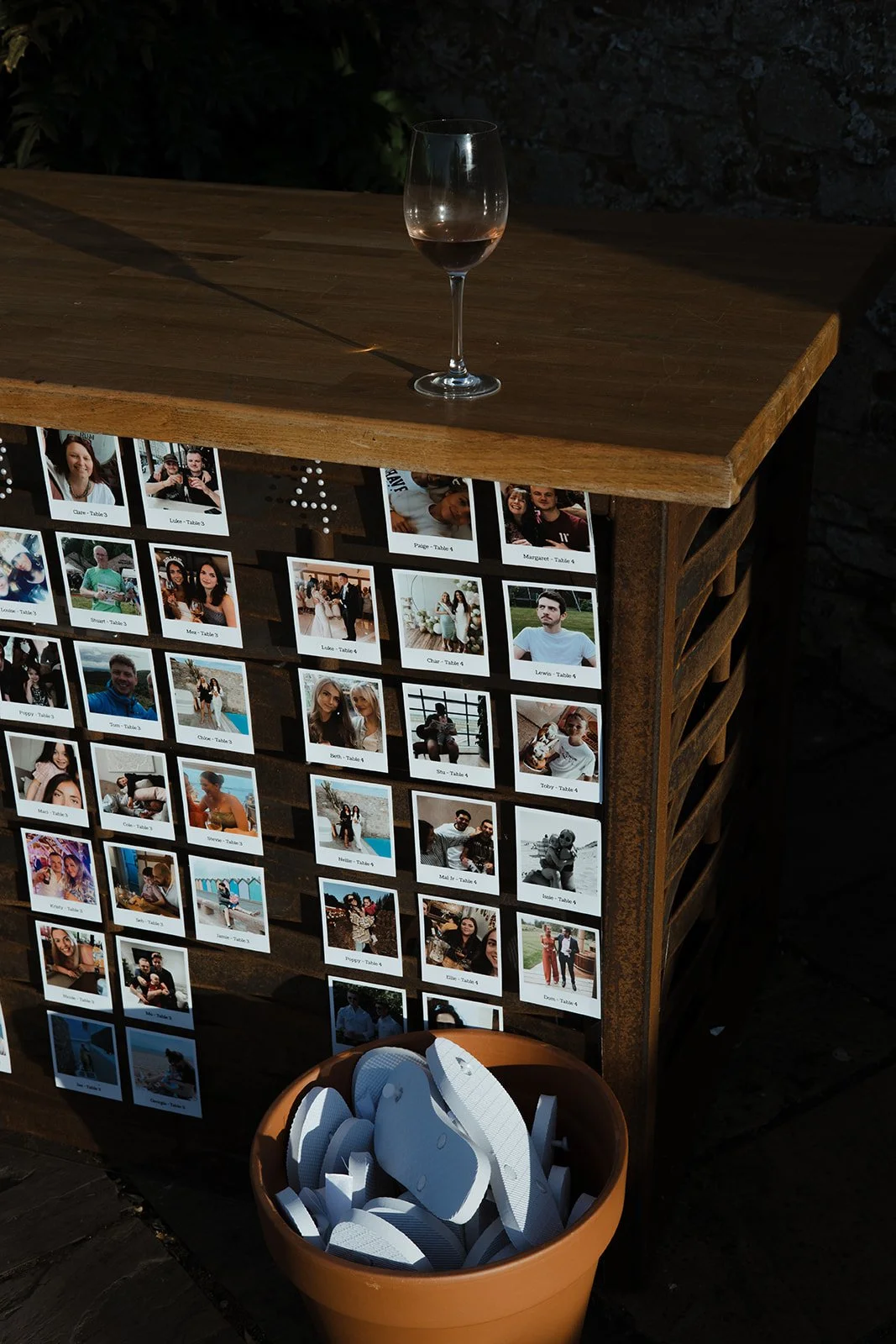 A wooden table with a glass of red wine on top, and a display of multiple printed photographs on the side of the table. A terracotta pot filled with white flip-flops is in the foreground.