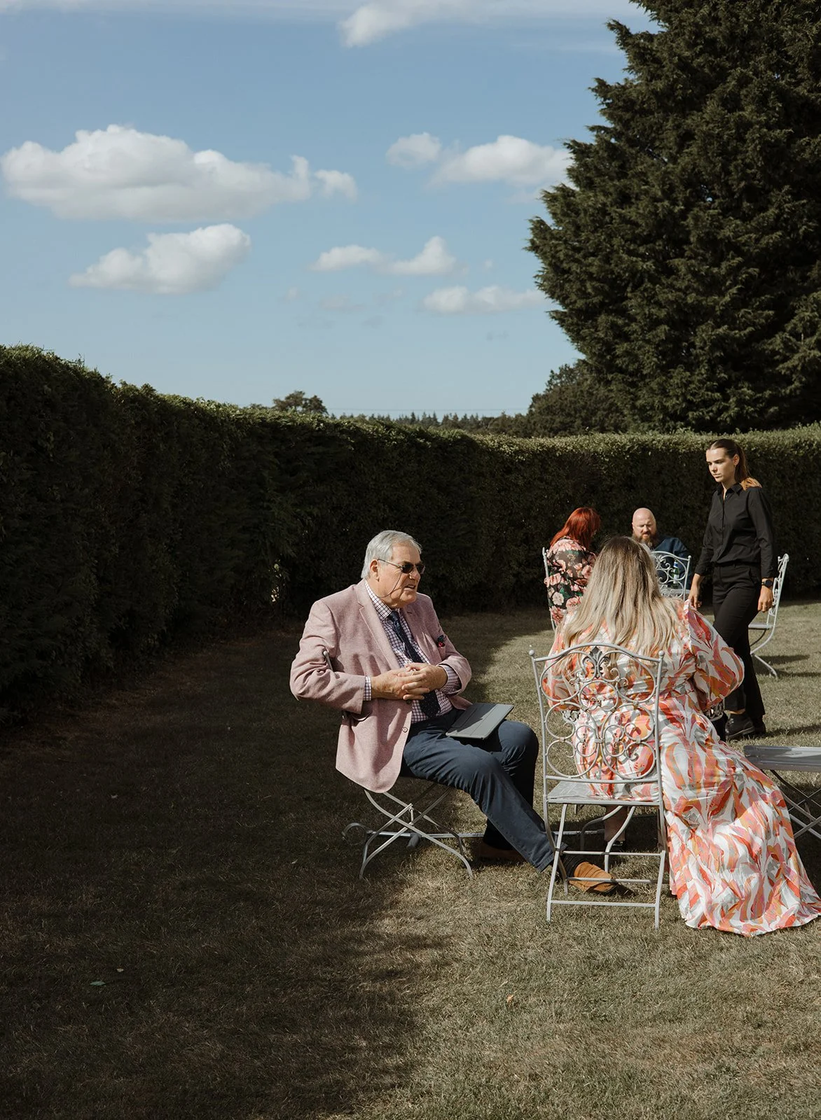 People sitting and standing in an outdoor garden setting, with a hedge and large trees in the background, under a partly cloudy sky.