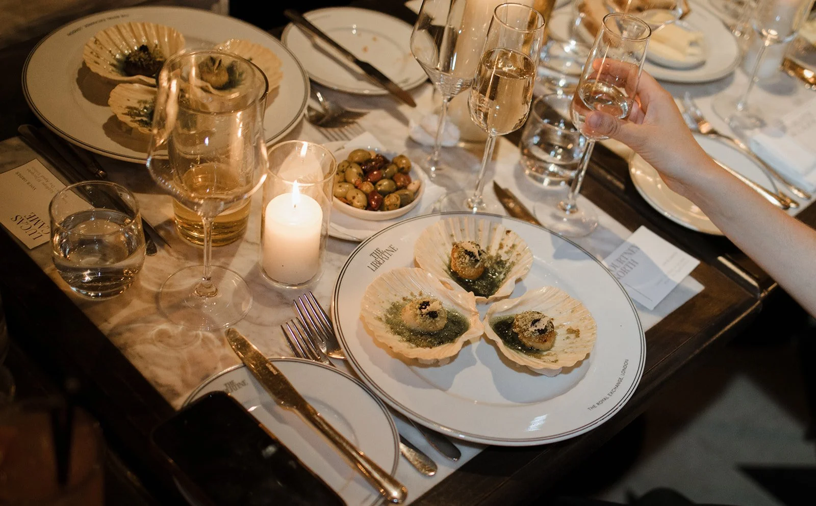 A fancy dinner table set with multiple glasses of water and champagne, plates with seafood dishes, a bowl of mixed olives, a candle, and silverware. A person's hand is holding a glass of champagne.