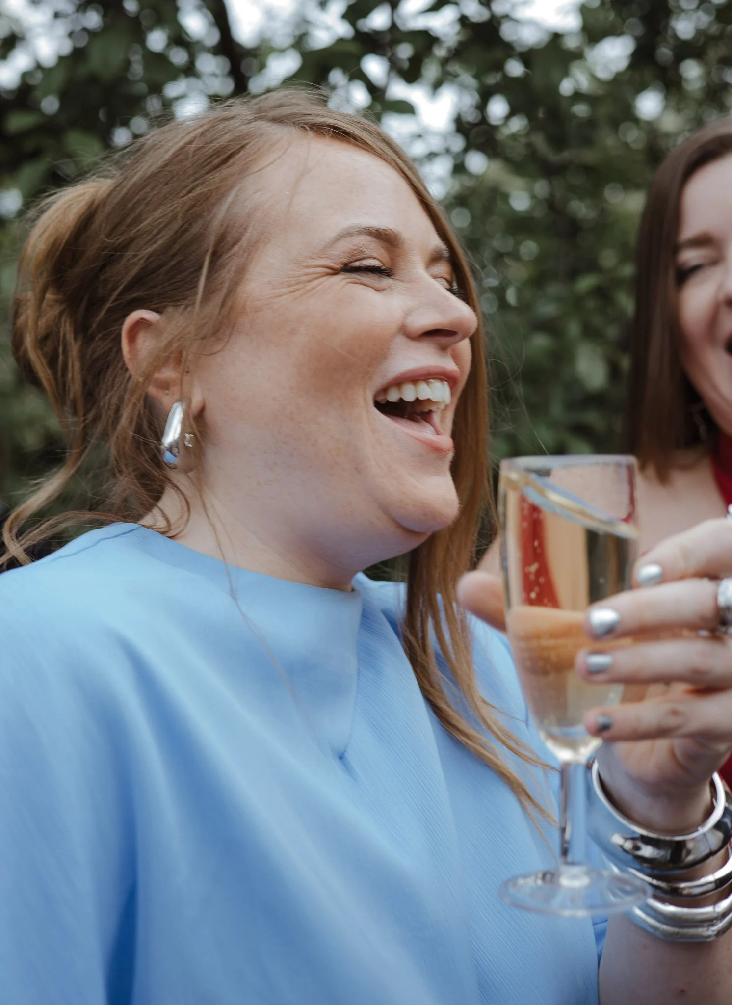 A woman wearing a blue top laughing and toasting with a glass of champagne outdoors, with another woman partially visible beside her.