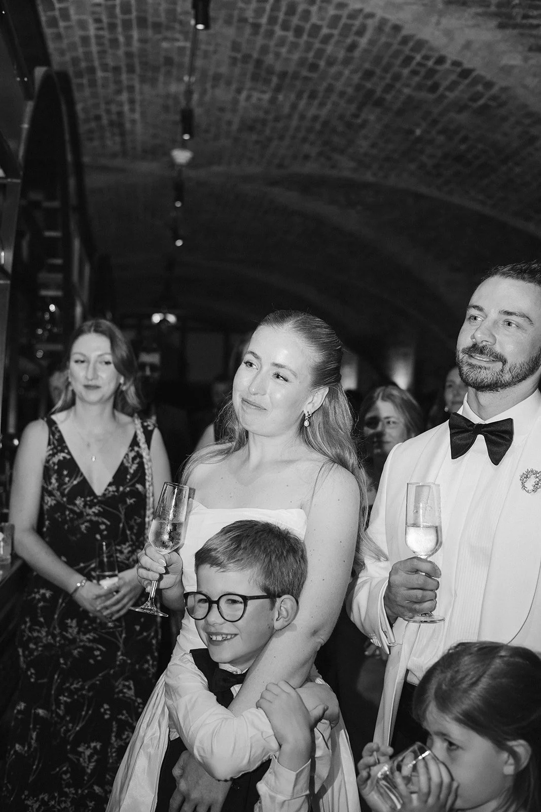Group of people at a formal event, including a woman holding a champagne glass, a man in a tuxedo holding a champagne glass, a boy with glasses smiling, and others in the background. The setting appears to be a bar or celebration venue with an arched